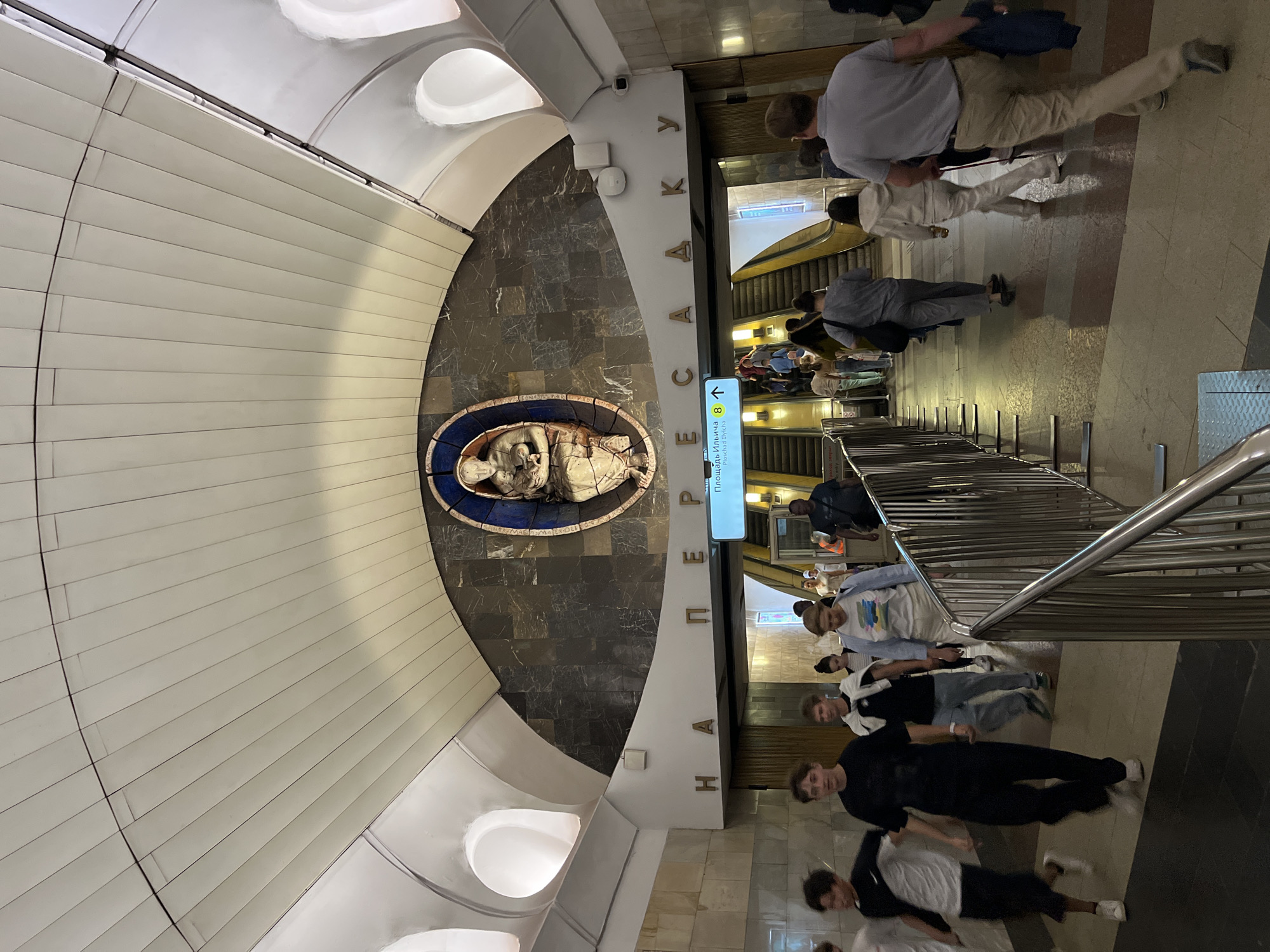 This is a photograph taken in the Moscow Metro, specifically at the Perervinskaya metro station, on the Lyublinsko-Dmitrovskaya Line. The scene depicts a bustling metro entrance area with multiple individuals moving in various directions.
In the foreground, several people are walking towards the viewer, some in groups and others individually. A group of young men, one in a black t-shirt and black pants, another in a black t-shirt and shorts with a white t-shirt over his shoulders, and a third in a black t-shirt and dark pants are visible on the left. They appear to be heading towards the escalators.
Further into the image, a long metal barrier guides the flow of people. Escalators are visible in the background, with a stream of commuters ascending and descending. A sign with Russian text, possibly indicating directions or station names, is mounted above the escalators. The architecture of the station is prominent, featuring a large, decorative mural above the entrance archway. The mural appears to be a carved relief of a figure holding a child, set within a circular, mosaic-like frame. The ceiling of the station is arched and lined with white panels, and the walls are clad in stone and marble. The overall impression is one of an active public transport hub.