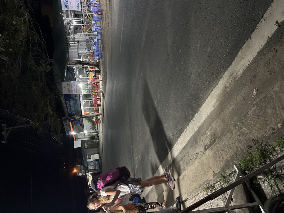 This nighttime image captures a street scene in Hà Tiên, Vietnam. Two individuals, appearing to be young women or girls, stand on a sidewalk in the foreground. One is wearing a white top and patterned shorts, carrying a prominent pink backpack, suggesting they might be travelers. The other is looking at a phone, dressed in a patterned top and shorts. A third person is partially visible at the bottom of the frame.

The setting is an urban street illuminated by artificial lights from surrounding businesses and streetlights. Along the sidewalk, several establishments, likely cafes or eateries, are visible with their lights on. Rows of blue and red plastic chairs are arranged outside these businesses for patrons. Overhead, power lines are visible against the dark sky, and some tree foliage is present on the left. The paved road next to the sidewalk features a distinct white line.

Visible text on signs confirms the location and type of businesses. "HÀ TIÊN" is clearly displayed, along with "CƯỜNG THỊNH" (which appears multiple times) and "BÃI GIỮ XE" (indicating a parking lot or bike storage). "CAFE VÕNG" (hammock cafe) is also partially legible. The time of day is clearly night, with the sky completely dark. The atmosphere appears calm, with no specific event other than people waiting or pausing their journey. A small white and red item, possibly a discarded can, lies on the pavement near the road.