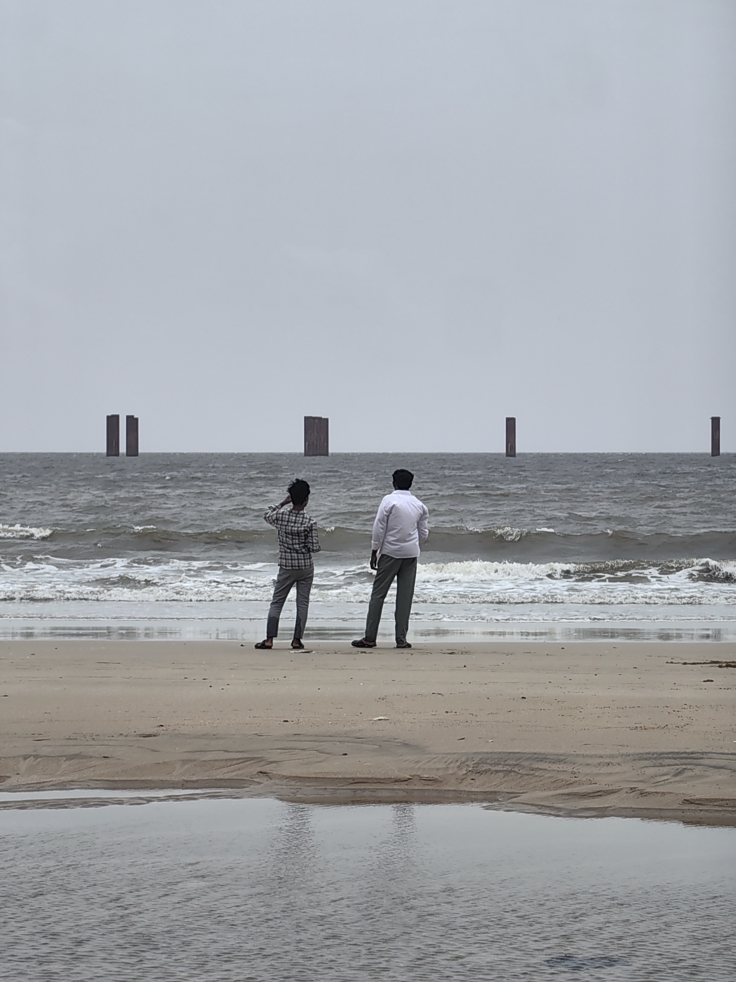 This image captures a desolate beach scene in Mumbai, India, under an overcast sky. In the foreground, a sandy beach is marked by shallow pools of water and rippled sand textures, suggesting the receding tide. Further out, the rough, grey waters of the Arabian Sea churn with moderate waves.
Standing on the shore, facing the ocean, are two young men. The individual on the left, dressed in a checkered shirt and grey trousers, is looking out towards the sea with his right hand touching the back of his head. The man on the right, wearing a white shirt and grey trousers, also faces the water, with his posture relaxed.
In the background, several tall, dark, rectangular structures rise vertically from the sea, spaced out across the horizon. These structures appear to be remnants of some construction or perhaps a jetty, adding a stark, almost abstract element to the otherwise natural seascape. The overall mood of the image is somber and quiet, with the muted colors and the presence of the solitary structures contributing to a sense of isolation.