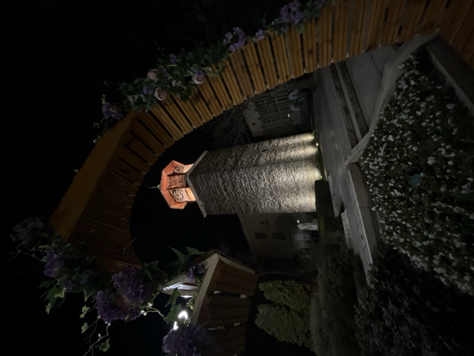 A nighttime, low-angle shot captures the illuminated Clock Tower in Sliven, Bulgaria, viewed through a decorative wooden archway. The central subject is a robust stone masonry tower, square in cross-section, with its lower portion brightly lit by at least three upward-facing spotlights. The upper section of the tower transitions to an octagonal wooden structure, featuring a reddish-brown wooden roof and a spire, with a clock face dimly visible on one side.
In the immediate foreground, a prominent curved archway, constructed from vertical wooden slats, is adorned with artificial purple floral arrangements and green foliage, including a hanging wooden planter box on the left. Below this archway, a flowerbed displays numerous small white flowers against dark foliage. To the left of the tower, evergreen shrubs and two conical evergreen trees are visible, alongside a multi-story building with light-colored walls and multiple windows. A white statue, appearing to be a human figure, stands in the landscaping between the building and the tower. Light-colored paved walkways are present, leading towards the base of the tower. To the right of the tower, a building with large windows is partially obscured by darkness. The background is a uniform dark sky, indicating night. No individuals or specific actions are depicted.