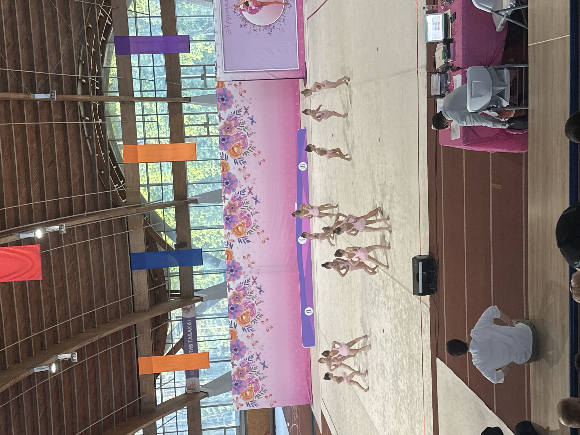The image shows a rhythmic gymnastics competition or performance taking place indoors, likely in Minsk, Belarus, as the banner in the background includes Cyrillic script. A group of young girls wearing pink leotards are performing on a light-colored floor mat. Behind them is a large, vertically oriented banner decorated with floral and butterfly patterns and a logo in the upper center. Colorful rectangular banners hang from the ceiling in red, orange, blue, and purple. To the right, there is a table with equipment, possibly for judges or officials, and a person is seated at the table. People appear to be watching the performance from the side. The building has a high, wooden ceiling and large windows providing natural light.