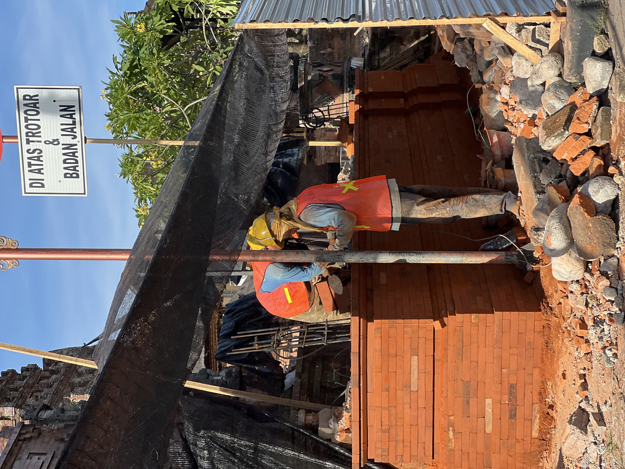 A construction worker in a yellow hard hat and orange vest is working on a brick structure under a makeshift scaffolding covered in black netting. Debris including bricks and stones is visible around the structure. A sign reading "DI ATAS TROTOAR & BADAN JALAN" is posted nearby. The location is the city of Denpasar, Indonesia.