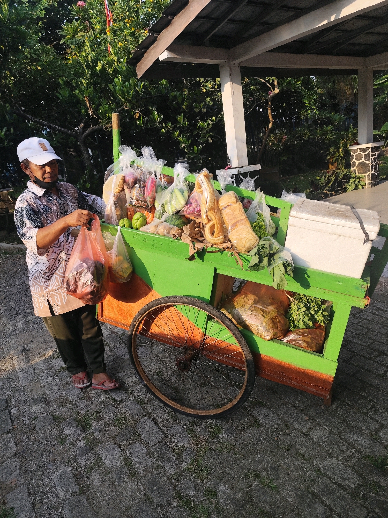 In Bogor, Indonesia, a person wearing a white cap and a patterned shirt stands next to a vibrant green and orange two-wheeled cart overflowing with produce. The cart is filled with various fruits and vegetables, many of which are bagged in clear plastic. Among the items visible are what appear to be cucumbers, peppers, leafy greens, and some packaged food items like crackers or bread. A white cooler box is also present on the top of the cart. The individual is holding a red plastic bag containing more food items. The scene is set outdoors, with lush green foliage and trees forming a backdrop. Sunlight casts shadows on the paved ground, suggesting it is either early morning or late afternoon. The cart's wheel is prominent, indicating it's a mobile vendor's setup.