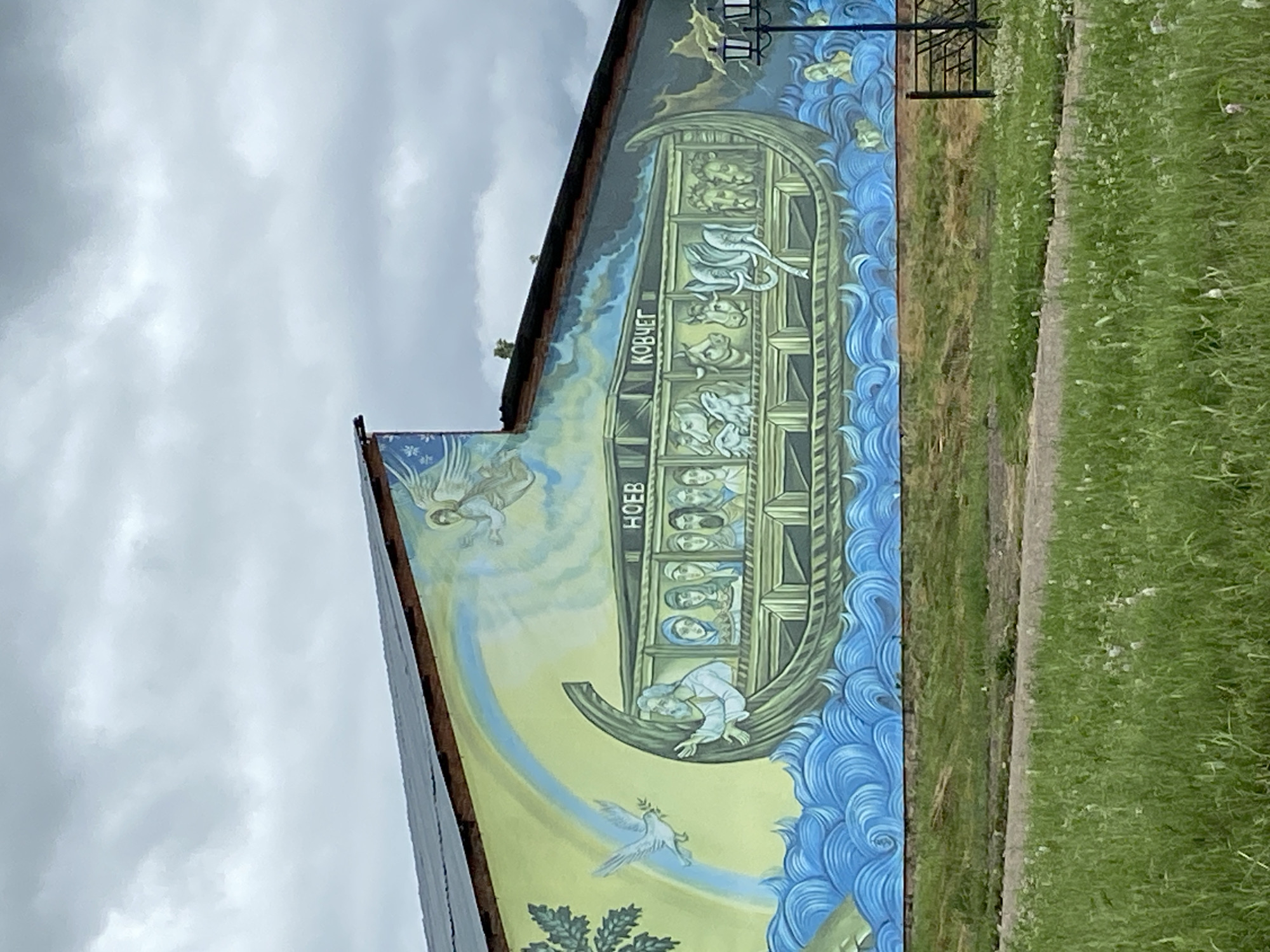 This is a photograph of a mural on the side of a building, depicting a scene from the biblical story of Noah's Ark. The mural, located in Кварса, Russia, shows a large ark with multiple levels filled with various animals, including what appear to be lions, elephants, and other creatures. Several people are visible inside the ark, looking out from different windows. An angelic figure is depicted in the upper left, and a dove carrying an olive branch flies in the lower part of the mural, symbolizing peace. The ark is surrounded by stylized blue waves. The mural is painted on a building that appears to be somewhat aged. To the right of the mural is a grassy area with a path. The sky above is cloudy, suggesting overcast weather. Text visible on the ark reads "КОВЧЕГ," which translates to "Ark" in Russian, and "НОЕВ," which translates to "Noah's."