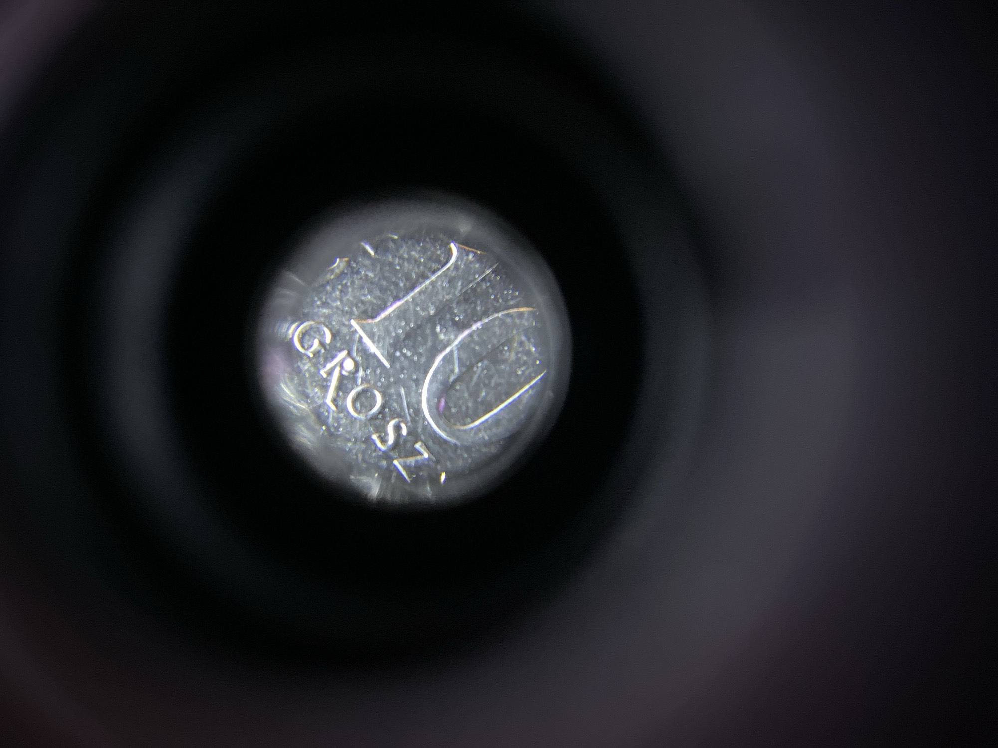 A close-up, macro photograph shows a portion of a coin, likely a 10 grosz coin from Poland, viewed through a dark, circular opening that resembles a lens or peephole. The coin is silver-colored and shows the number "10" prominently, along with the inscription "GROSZ". The surface of the coin exhibits a textured, slightly worn appearance with visible scratches and imperfections, indicating it has been in circulation. The surrounding darkness is out of focus, creating a bokeh effect that isolates the coin as the primary subject. The lighting suggests a controlled environment, possibly a studio or laboratory setting, rather than an outdoor or urban scene. There is no discernible information or context that would link this image to the city of Zgrade, Montenegro.