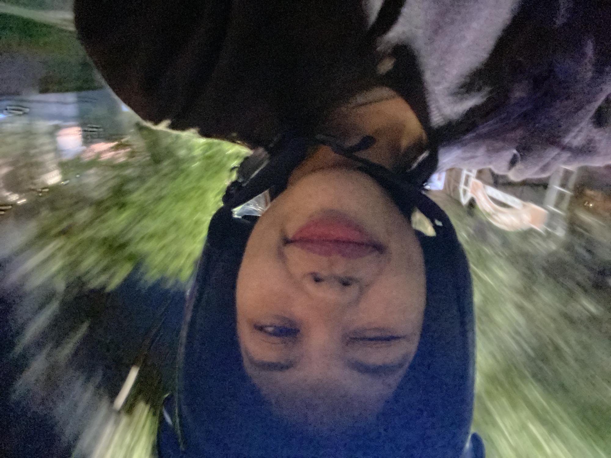 The image is a selfie taken by a young woman wearing a black helmet. She is on a motorbike, as indicated by the motion blur of the background which shows trees and streetlights. The woman is looking at the camera and winking, with her mouth slightly open as if she is smiling. She is wearing a dark-colored jacket with lighter gray accents. The background is blurry and shows a street at night with a pinkish hue in the distance, possibly from neon lights or sunset. The overall impression is of a playful and candid moment captured while on the move. The location appears to be a street in Madiun, East Java, given the context.