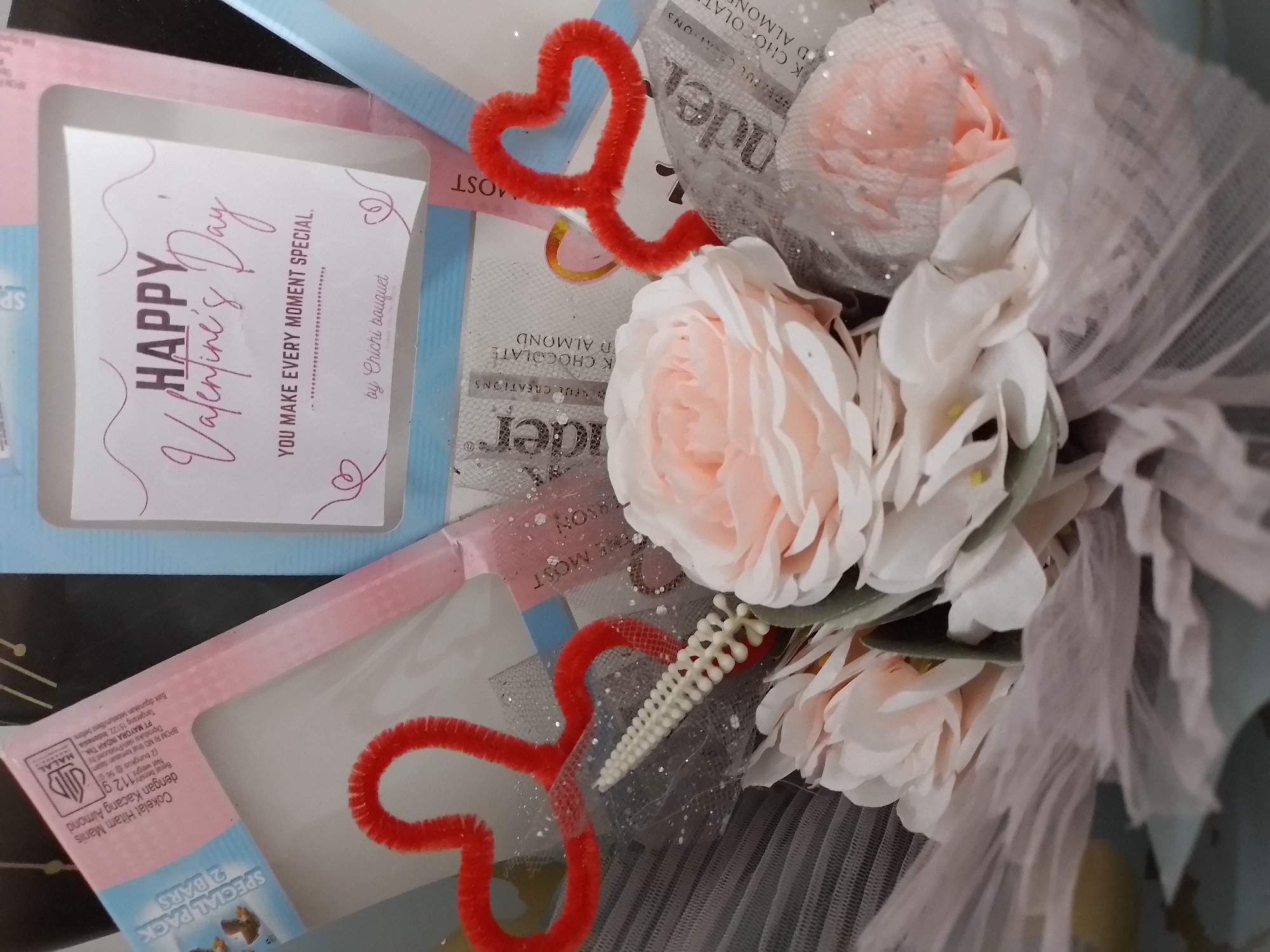 The image shows a close-up of a Valentine's Day gift arrangement. The arrangement includes faux flowers in shades of pink and white, red pipe cleaner hearts, and individually wrapped chocolate bars. A card with "HAPPY Valentine's Day" and "YOU MAKE EVERY MOMENT SPECIAL" is visible. The gift is adorned with sheer, pale pink fabric. The background is blurred, but suggests an indoor setting with some dark surfaces and hints of blue and pink packaging. There are no people or specific city landmarks visible, therefore no location in Ngancar, Indonesia can be identified.
