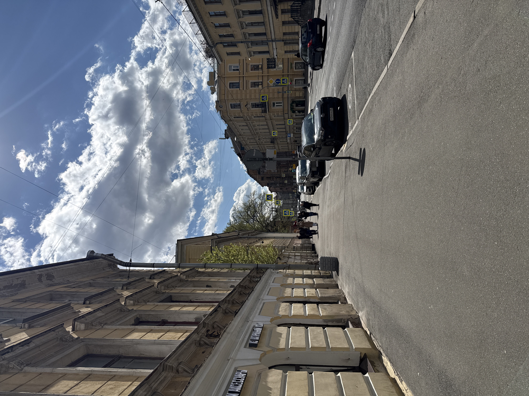 The image depicts a street scene in Saint Petersburg, Russia. The perspective is from the sidewalk looking down the street. Buildings line both sides of the road. Parked cars are visible along the right side of the street. Several people are walking on the sidewalk further down the street near a pedestrian crosswalk. The sky is visible above the buildings, with blue patches and white clouds. Overhead wires crisscross the sky. The architecture of the buildings is characteristic of Saint Petersburg. Signage in Russian is affixed to the building on the left.