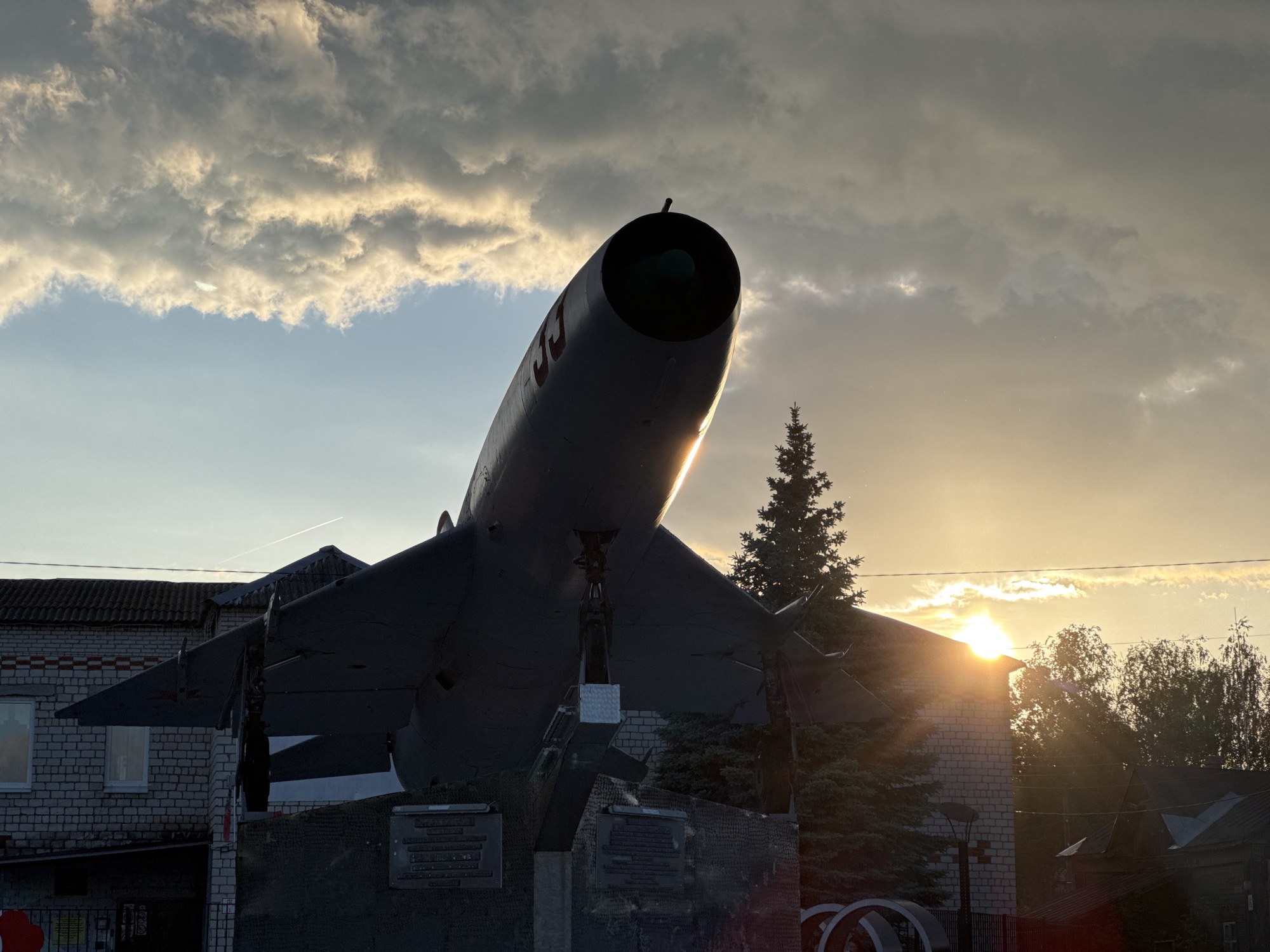A large, dark aircraft, likely a fighter jet or reconnaissance plane, is displayed as a monument, viewed from a low angle, silhouetted against a dramatic sunset sky. The aircraft features a long, cylindrical fuselage with a prominent nose cone and swept-back wings. Red Cyrillic letters, possibly "CCCP" (USSR), are visible on the side of its fuselage. It is mounted on a substantial, dark-colored pedestal which appears to have memorial plaques, though their text is indistinct.
The scene unfolds at sunset or late afternoon, with the sun low on the horizon, casting intense backlighting and creating a strong lens flare near the right side of the image. The sky is filled with a mix of clouds, some brightly illuminated by the setting sun, others appearing darker and more voluminous. A tall, dark coniferous tree stands to the right of the aircraft, also partially silhouetted. In the background, there are brick buildings with tiled roofs and windows, adding to the urban or town setting. No people are visible. The overall impression is one of a static outdoor display under a striking sky.