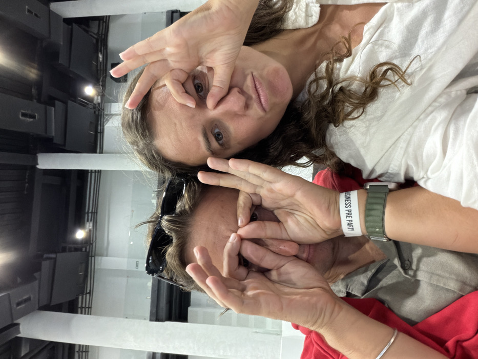 Two women pose close together, making circles around their eyes with their fingers. The woman on top wears a white shirt and has wavy hair. The woman below wears sunglasses perched on her head and a red shirt. A white wristband with "BUSINESS PRE PARTY" is visible on her wrist next to a green watch band. The background shows interior architecture with columns and dark panels. The image is oriented sideways.