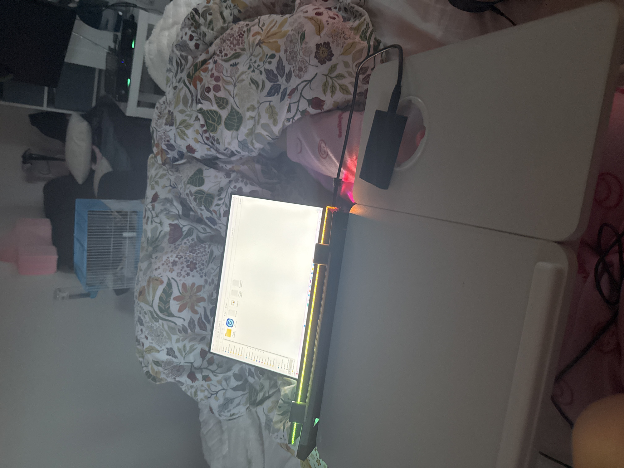The scene depicts an indoor residential environment, specifically a dimly lit room, likely a bedroom, in the City of Zagreb, Croatia. The exact address or specific location within Zagreb is not discernible from the image.
In the foreground, a light-colored portable bed desk is positioned over a bed. A laptop with an illuminated screen displaying a file explorer window (showing "Documents," "Pictures," and other folders) rests on the left side of this desk. The laptop features a multi-colored LED light strip at its base, below the screen. A black, rectangular electronic device, possibly a USB hub or external storage, is connected via a black cable and rests on the right side of the desk, partially within a circular cutout, emitting a red light.
The bed is covered with a patterned duvet featuring botanical designs in various shades of green, orange, red, purple, and yellow on a light background. White bedding is visible beneath the duvet. A person's lower leg or knee, covered by pink fabric, is partially visible in the bottom left corner beneath the desk. Additional pink fabric, possibly clothing or a blanket, is visible beneath the electronic device on the desk.
In the mid-ground, to the left, a light grey wall is visible. A small blue pet cage with white bars is present, with a pink object positioned above or behind it. To the right, a white shelving unit or piece of furniture holds several electronic devices, some exhibiting green indicator lights. A dark, rectangular object, possibly a television or monitor, is atop this shelving unit. A light-colored pillow rests on a dark upholstered furniture piece, likely a chair or additional bedding, to the right of the pet cage.
The room is generally dim, with the laptop screen and its base lighting serving as the primary light sources in the immediate area.