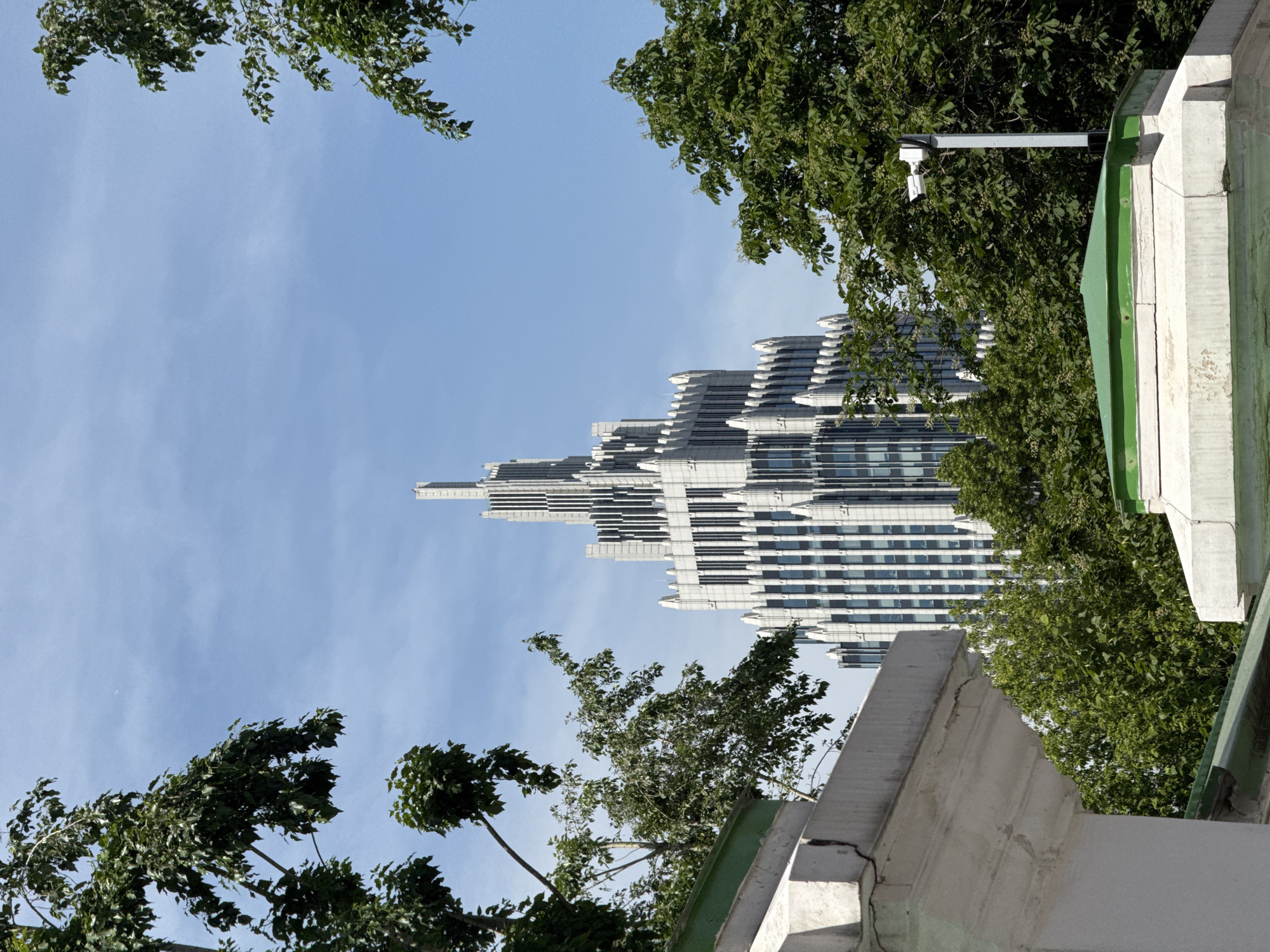 The scene depicts the Triumph Palace skyscraper in Moscow, Russia, prominently featured under a clear blue sky with minimal high-altitude clouds. The building, constructed in a neo-Stalinist style, presents a light-grey, multi-tiered, stepped-back facade, culminating in a slender central spire. Vertical window lines punctuate the building's exterior. Lush green deciduous trees and dense foliage frame the skyscraper on the left and right sides, partially obscuring its lower levels. In the foreground, white concrete architectural elements, possibly walls or parapets, are visible, topped with weathered green sections. A white security camera on a grey pole is positioned on the right side, partially visible above one of the concrete structures and amidst the foliage. No individuals are present in the image.