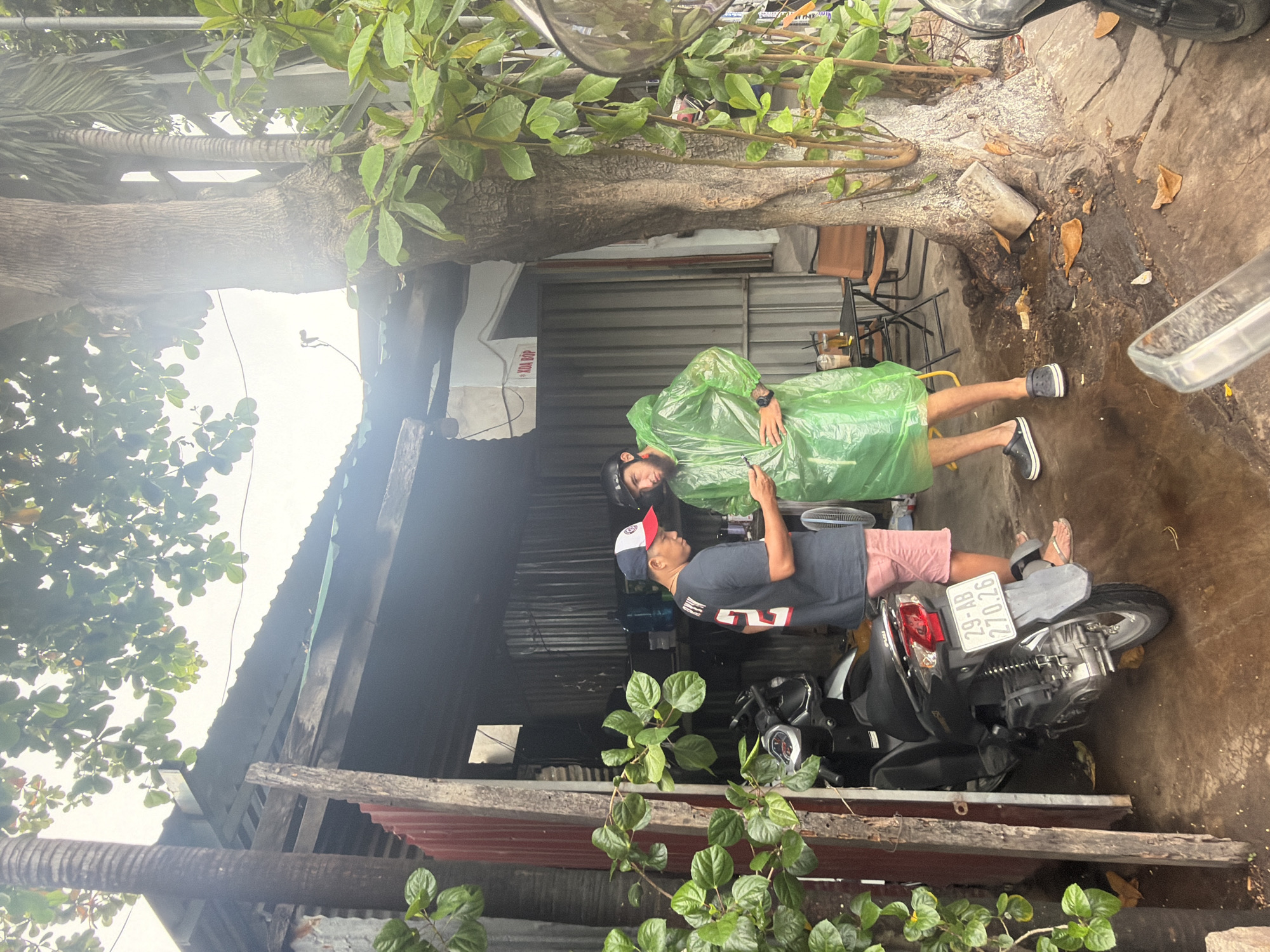 The image depicts an outdoor scene in Nha Trang, Vietnam. Two men are engaged in conversation near a building with a corrugated metal exterior and a tin roof. The man on the left, wearing a dark t-shirt with "RYN" and a number "24" on the back, and pink shorts, is looking at a smartphone. He has a baseball cap on. The man on the right, wearing a bright green poncho and a helmet, is also looking at the smartphone. A motorcycle is parked to the left of the building. The ground is wet, suggesting recent rain. A large tree with dense foliage stands to the right of the building, and a palm tree is visible behind it. There are tables and chairs set up near the tree, suggesting an outdoor seating area. The overall impression is a street scene in a tropical climate.