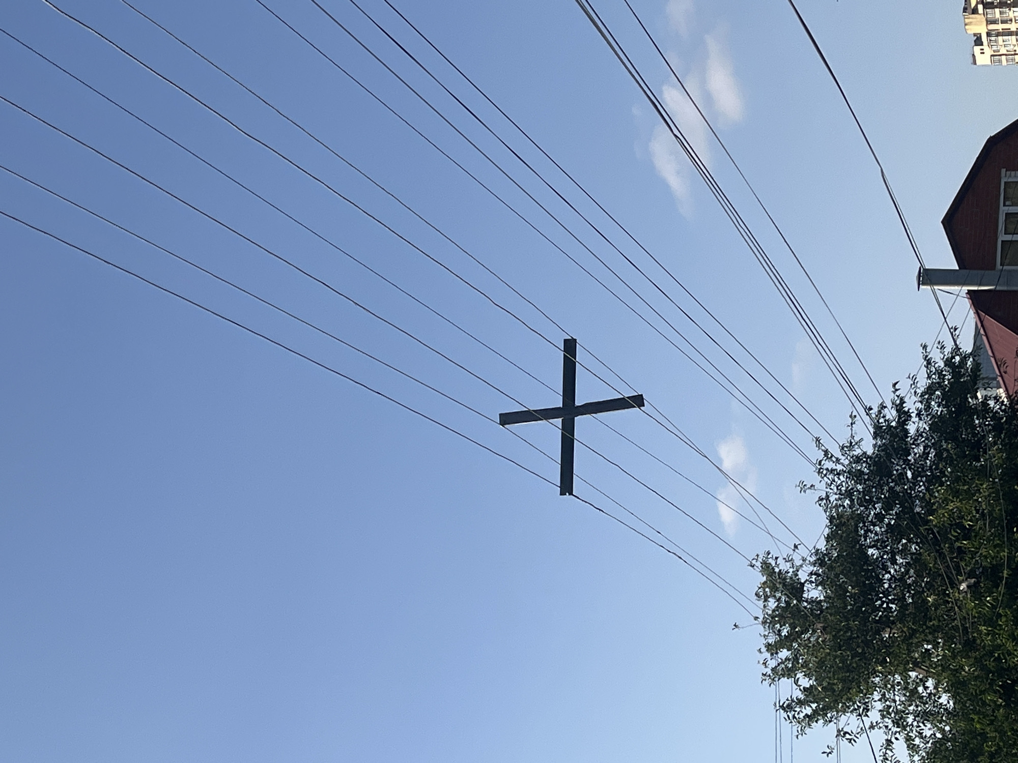 The image shows a view of power lines against a blue sky. A black metal structure in the shape of a plus sign is suspended from the lines. A tree with green foliage is visible on the right side of the frame, obscuring part of a building. The top of another building can be seen in the upper right corner. The photo was taken in Krasnodar, Russia.