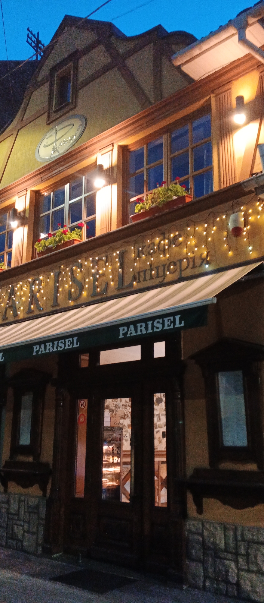 The image depicts the exterior of a building at dusk, likely a restaurant or cafe, with the name "PARISEL" prominently displayed. The building features a traditional architectural style with a yellow facade, dark wooden trim, and a mansard roof with exposed beams. Small potted plants with red flowers are placed on a ledge above the windows. The entrance is marked by a dark wooden door with glass panes, and above it is an awning also displaying the "PARISEL" name. Strings of warm white lights are draped across the upper facade, illuminating the signage and the building's details. The overall impression is that of a cozy and inviting establishment. The location is identified as Berehove, Ukraine.