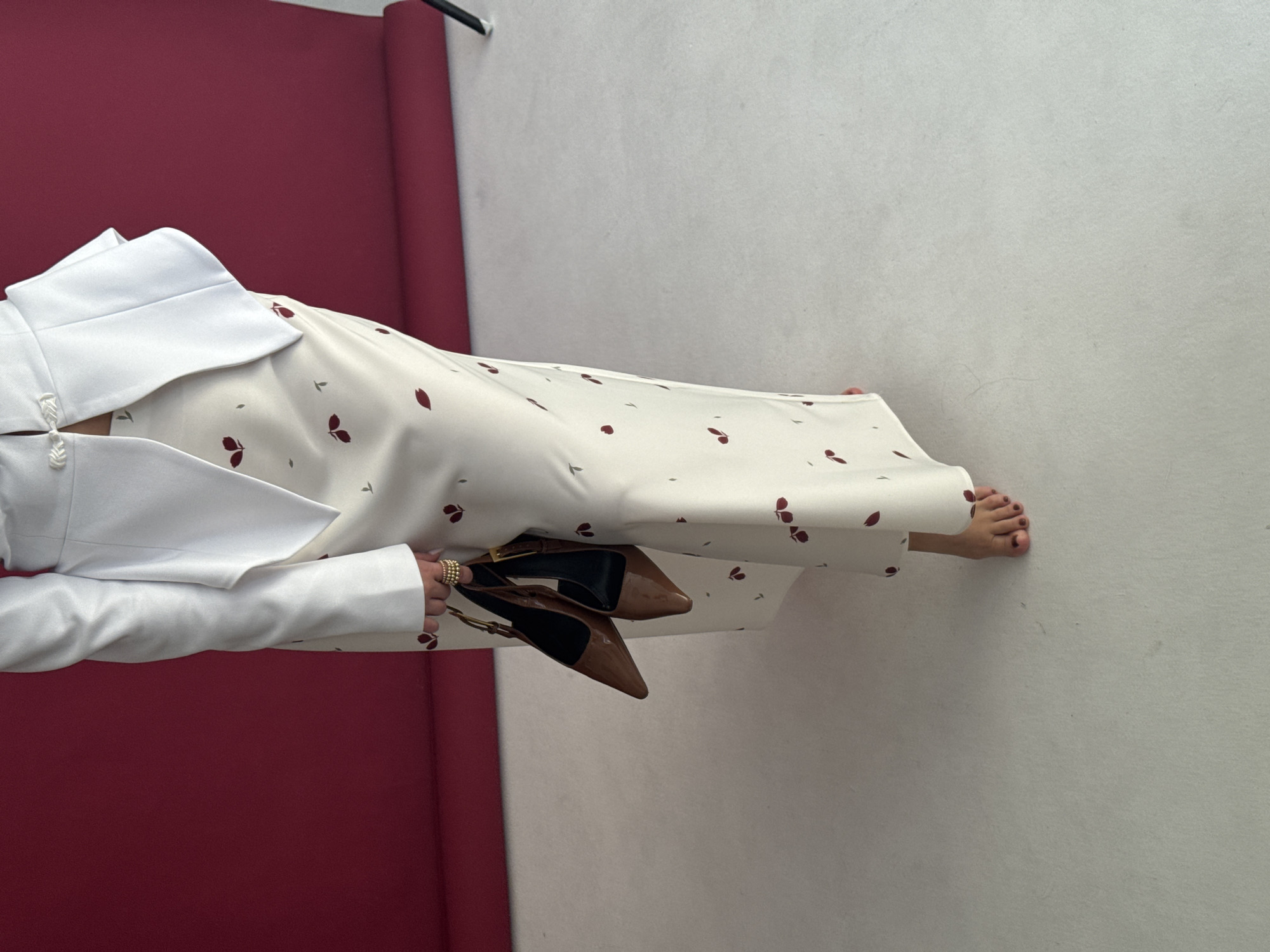 The scene depicts a partial view of an individual within what appears to be a photography studio, identified as being in Chaikovsky, Russia. The subject is visible from the waist down, standing on a seamless white floor. They are wearing a white, structured jacket with an asymmetrical hem and a distinctive frog closure on the upper left side, over a cream-colored, floor-length skirt or dress patterned with scattered dark red leaf or petal motifs. The person is barefoot, with toenails painted a dark red hue. In their right hand, they hold two brown patent leather slingback high-heel shoes by their straps. Multiple gold-toned rings are worn on the fingers of the right hand. The background is a seamless deep red photography backdrop, which curves from the vertical plane to meet the white floor. A portion of a black stand is visible on the far right edge of the frame against the red backdrop.