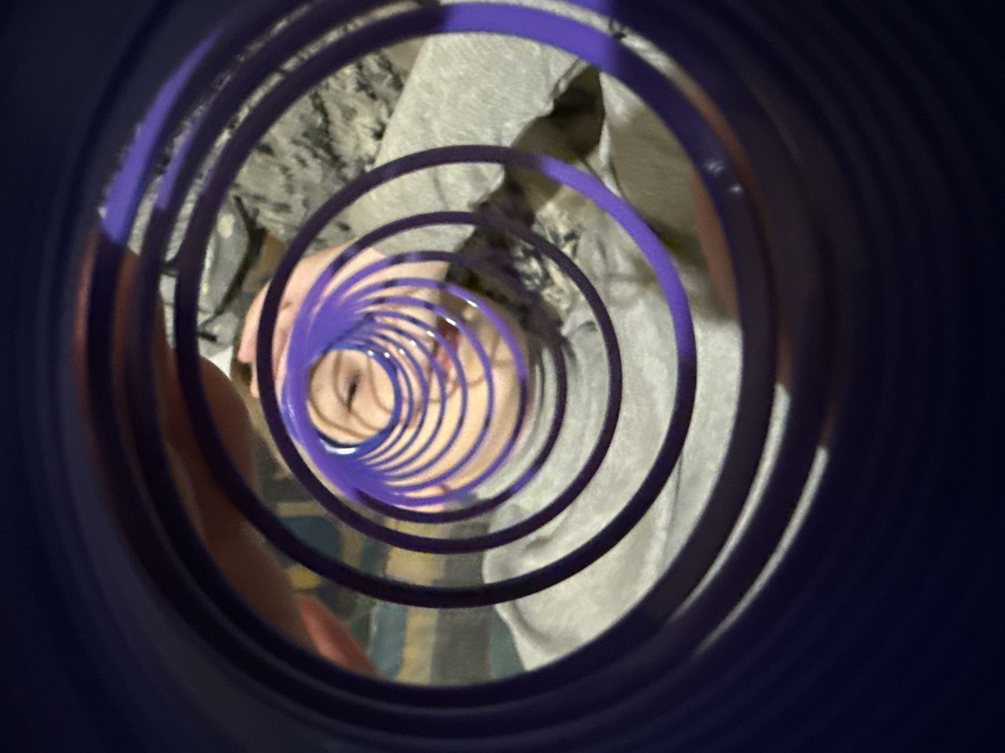A child's face is visible at the center of a purple, spiral-shaped play structure. The background includes a light gray surface, possibly clothing or a playground feature. The scene is viewed from inside the play structure. There is no information to identify the city of Dmitrov, Russia, in the image.