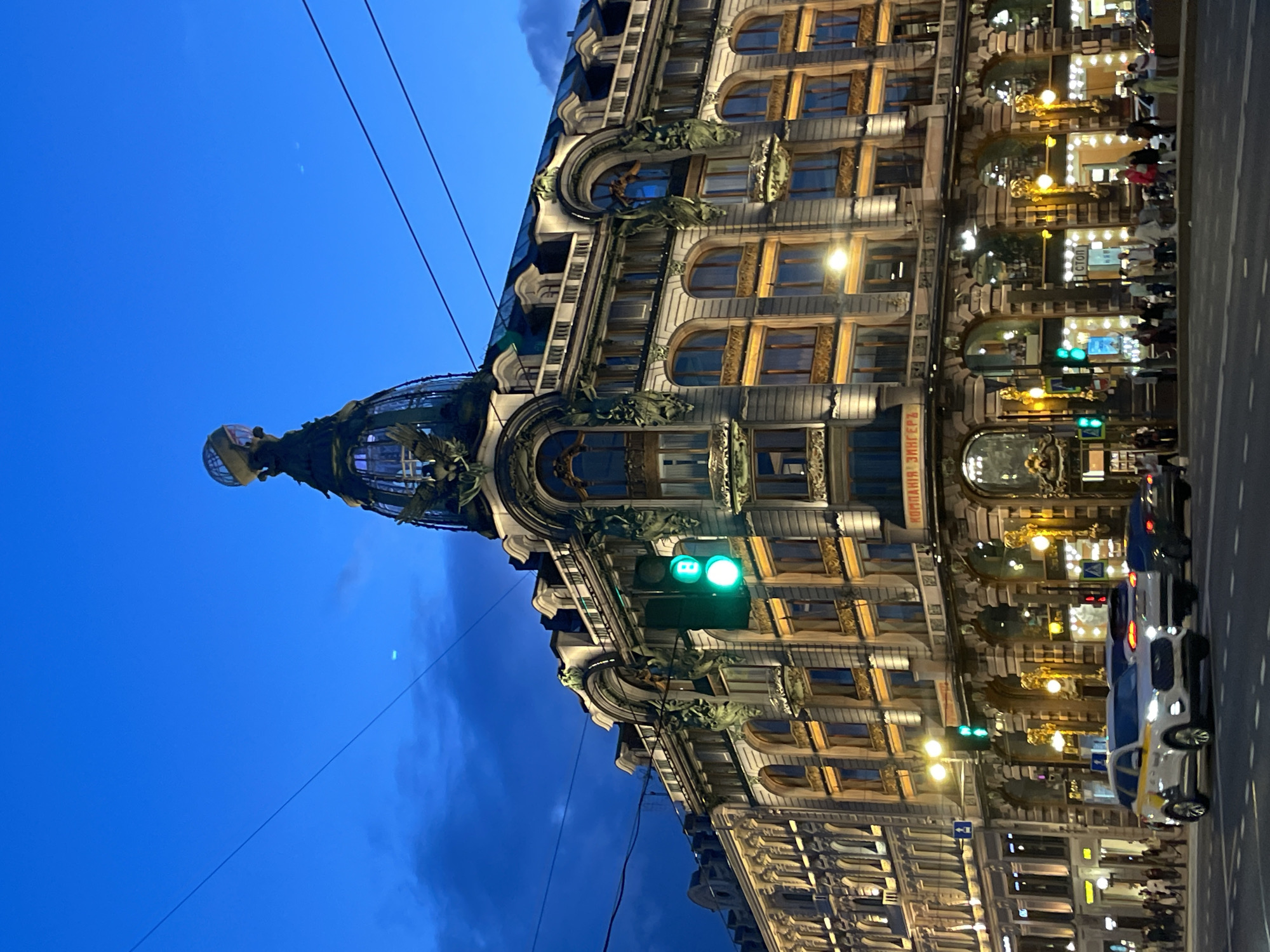 This media file depicts an evening urban scene in Saint Petersburg, Russia, focusing on the ornate architecture of a prominent building, likely the Singer House (Dom Knigi). The multi-story building occupies the left side of the frame, showcasing a richly decorated facade with classical elements, numerous arched windows, and a distinctive glass dome or cupola at its corner. Many windows are brightly illuminated, suggesting interior lights from businesses or residences.
The sky is a deep, rich blue, indicating twilight or nightfall, with some darker cloud formations. Overhead wires or cables stretch across the upper part of the image. On the right, a city street with white lane markings is visible, populated by several vehicles, including a white car with blue and yellow markings, possibly a police car or taxi. Multiple traffic lights are visible, predominantly displaying a green signal. Faint figures of people can be discerned within the brightly lit ground-floor windows and possibly on the sidewalk.
Visible text in Cyrillic includes "СТОП" (STOP) on a sign within one of the ground-floor windows, and "КОМПАНІЯ" (COMPANY) partially visible on another sign. The scene conveys a lively urban atmosphere after dark.