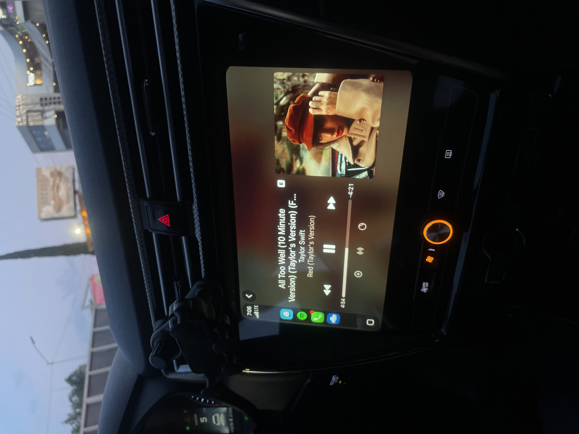 The image shows the dashboard of a car, displaying a song playing on the infotainment system. The song is "All Too Well (10 Minute Version) (Taylor's Version) (F...)" by Taylor Swift. The car's air vents, hazard lights button, and other dashboard elements are visible. A phone mount is attached to the air vent. The time displayed on the phone is 7:06. Outside the window, city elements of Puebla, Mexico, are visible in the background.