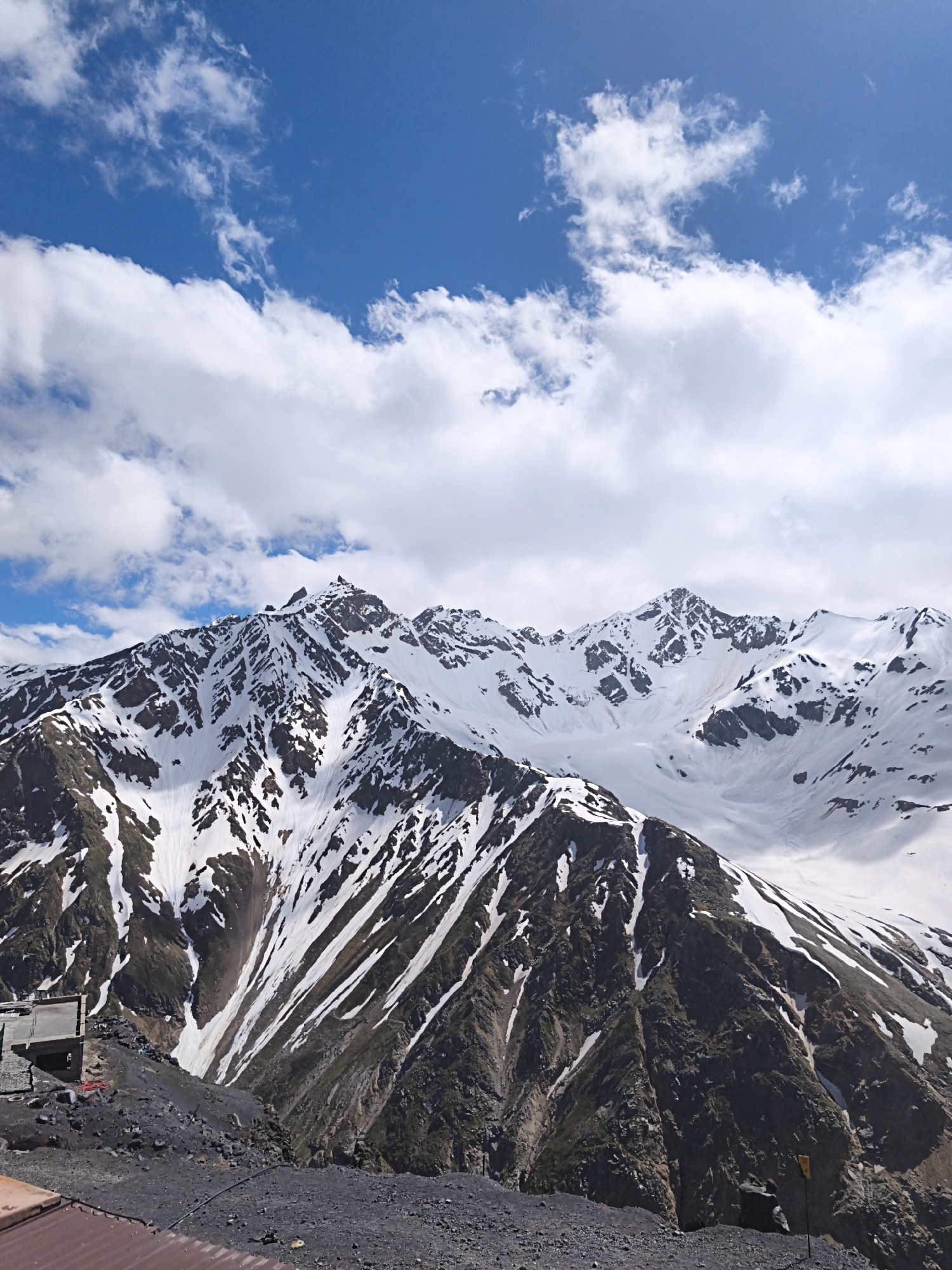 The image depicts a mountainous landscape in the vicinity of Terskol, Russia, under a partly cloudy blue sky. In the foreground, there is a rocky, dark gray ground with some scattered debris and a section of a corrugated metal roof. Further back, a concrete structure, possibly a small building or platform, is visible on the left side of the frame.
The dominant feature of the image is a large, snow-capped mountain range. The mountains are rugged, with dark rocky slopes interspersed with patches of white snow. The snow cover is uneven, suggesting the effects of melting or wind erosion, creating a dramatic contrast between the dark rock and the bright snow. Streaks of snow run down the mountainsides in various directions. The peaks are sharp and jagged, indicative of a high-altitude environment.
In the sky, scattered white clouds are present, with areas of clear blue sky visible between them. The lighting suggests it is daytime, with the sun illuminating the scene. There are no people or animals visible in the image. The overall scene conveys a sense of natural grandeur and the harsh beauty of a high mountain environment.