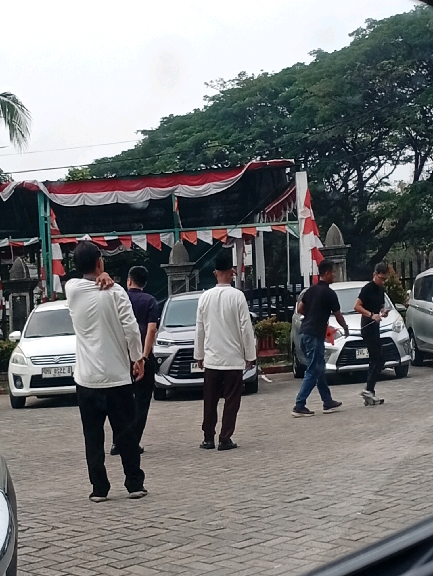 The image depicts an outdoor scene with several individuals and vehicles, likely in the city of Kelapa Dua, Indonesia. In the foreground, a person wearing a white long-sleeved shirt and black pants stands with their back to the camera, touching their right shoulder. To their left, a white car is parked. Further back, a person in a dark shirt and black pants walks away from the camera, followed by a person in a black shirt and blue jeans who appears to be gesturing with their hands. To the right, a silver car is parked, and two individuals are visible near it. One is wearing a black shirt and black pants, and the other, partially obscured, is wearing a white long-sleeved shirt and maroon pants. Behind the people and cars, a decorative canopy with red and white banners is visible, suggesting a potential event or celebration. Tall trees with dense green foliage form the background, and the ground is paved with interlocking brick tiles. The sky is overcast.