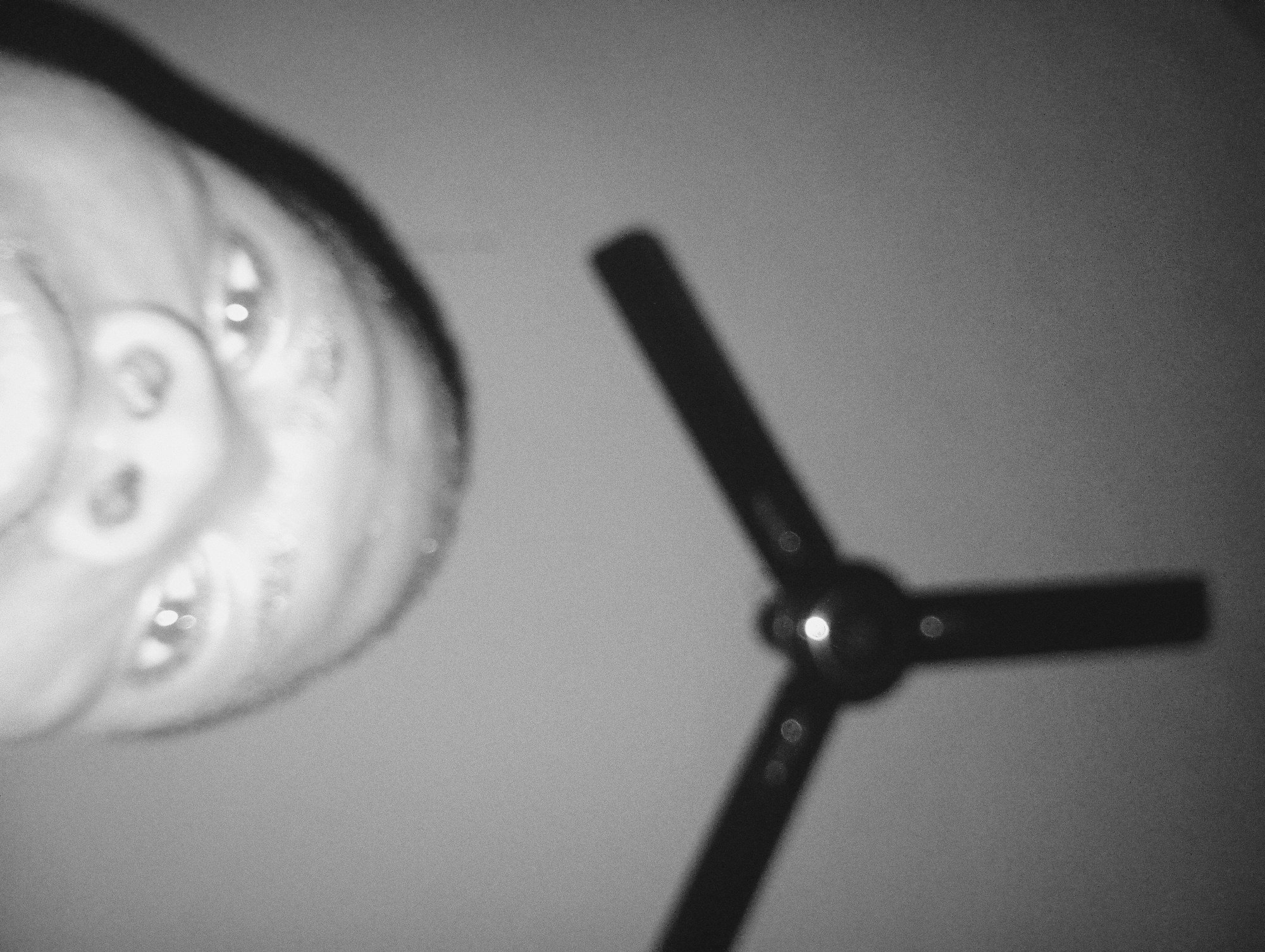 A grayscale image shows an upward-facing human face on the left and a blurred three-pronged object on the right, against a uniformly lit background. The face appears to be close to the camera, with visible facial features including eyes, nose, and the edge of hair. The three-pronged object, possibly a ceiling fan or a similar device with three blades, is out of focus. No specific details indicate that the location is Nkpor, Nigeria; this cannot be determined from the image. There are no visible actions or interactions.