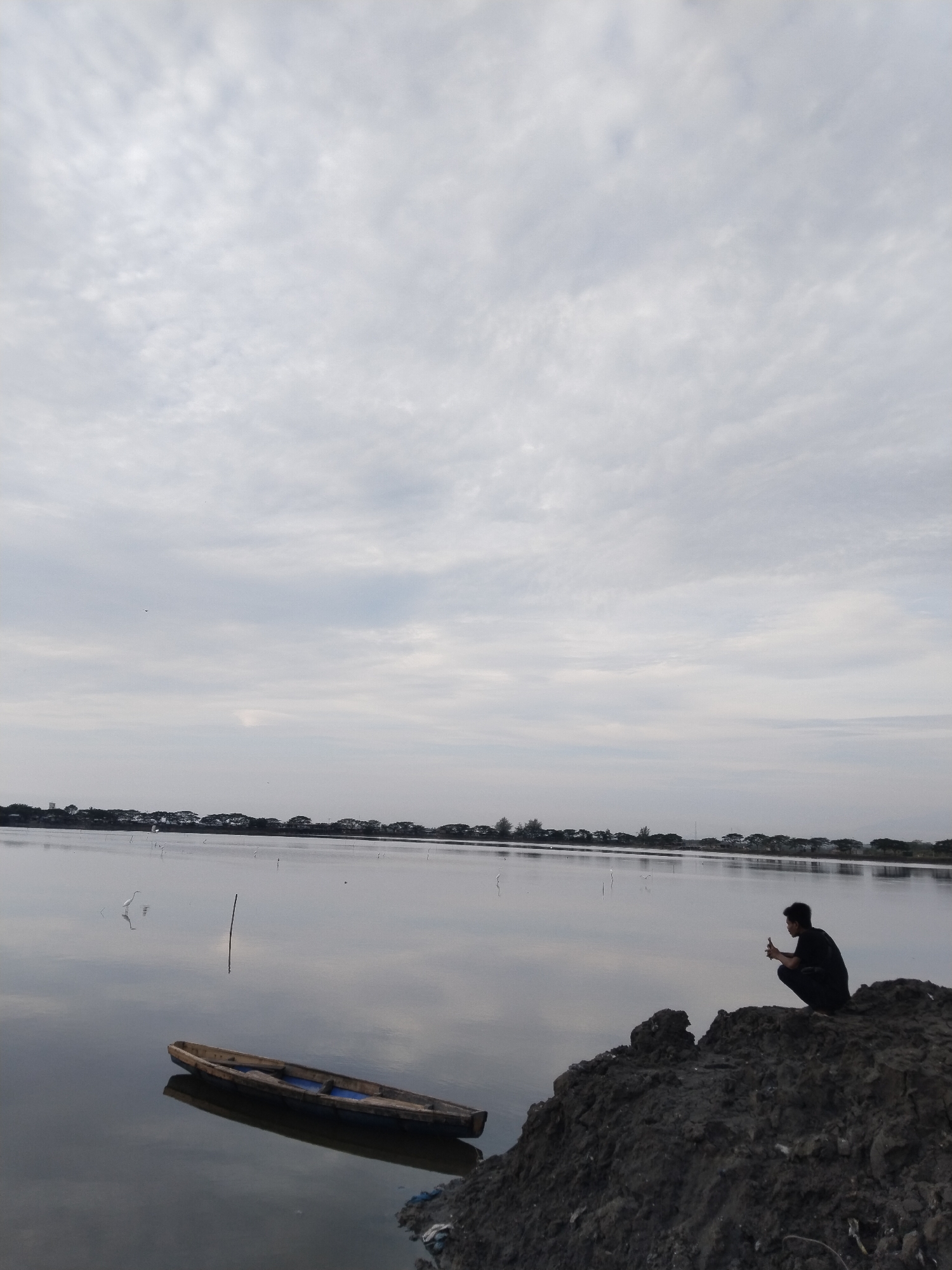 A person is crouching on a mound of dark soil overlooking a calm body of water. The person, wearing a dark shirt and pants, is holding a cell phone to their ear with their right hand, as if making a call or taking a photo. To the left of the mound, a long, wooden boat with blue seating is partially submerged in the water, its reflection clearly visible on the surface. The water stretches out to the horizon, where a line of trees and what appear to be buildings are silhouetted against a cloudy sky. Several white birds, possibly egrets, are visible in the water and in flight. The overall atmosphere is tranquil and overcast. The scene is likely located in Lhokseumawe, Indonesia, given the typical landscape of aquaculture ponds and coastal environments found in the region, which often feature such boats and open water.