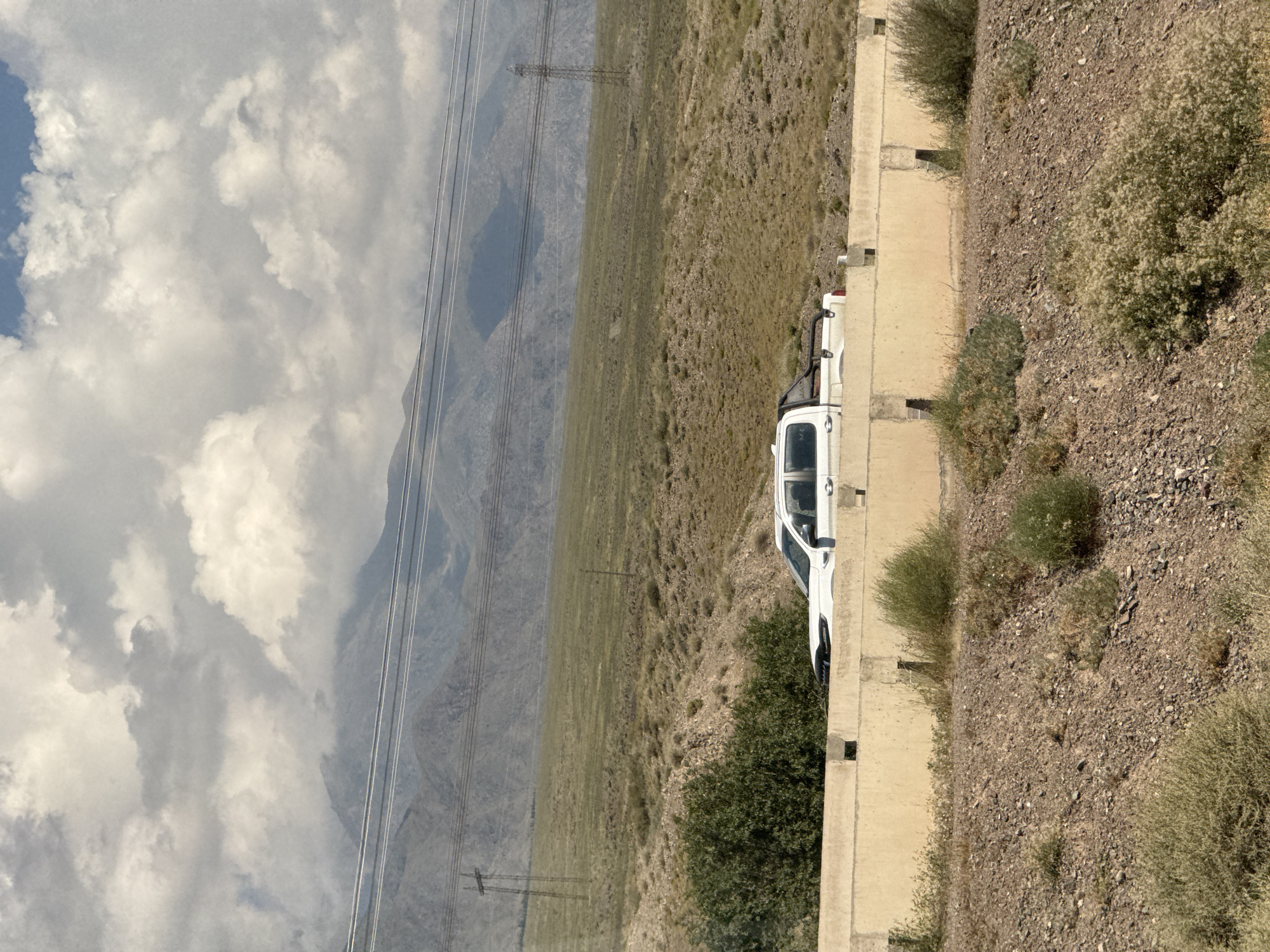 A white pickup truck is parked behind a concrete barrier on a dirt road. The truck is facing right and has its headlights on. The truck is in a dry, grassy area with mountains in the background. The sky is cloudy. The location is Kok-Moinok - 1, Kyrgyzstan.