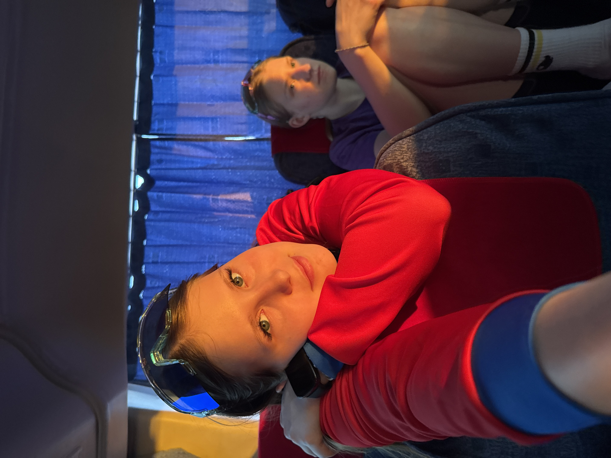 The image shows two young girls on a bus. The girl in the foreground is wearing a red sweatshirt with blue accents and sunglasses on her head. She is holding a phone to her ear and looking at the camera. The girl in the background is wearing a purple shirt, socks with dark accents, and has a headband on. Blue curtains hang behind them. The location is not evident from this image, and cannot be assumed to be Osa, Russia.