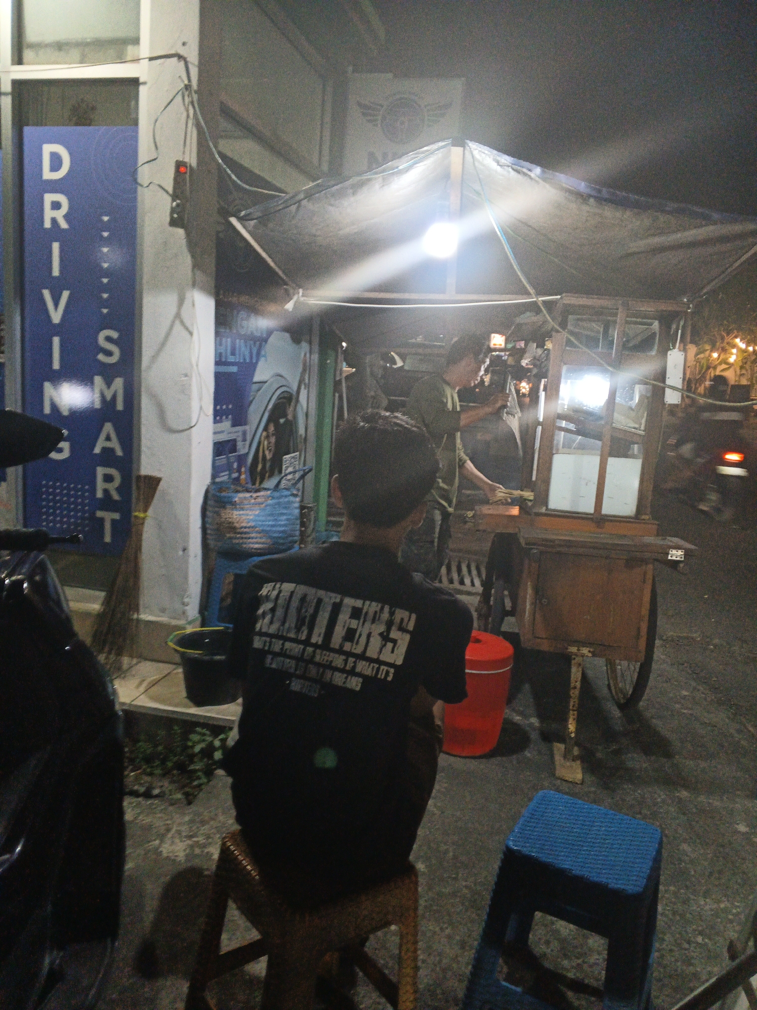 This is a nighttime photograph of a street scene in Sukawati, Indonesia, with a focus on a food cart. A young man in a black t-shirt with the word "Hunters" printed on it in white, is seated on a wooden stool facing away from the camera. He appears to be a customer. Behind him, a food vendor, also a male, is operating a wheeled food cart. The cart has a wooden frame and glass display cases. The vendor is seen reaching into the cart, possibly preparing food. Several items are visible on and around the cart, including what appear to be food items, utensils, and cleaning supplies. In the background, there are buildings, streetlights, and other elements of an urban environment. A blue sign with white text reading "DRIVING SMART" is visible on the left side of the image. The overall lighting suggests it is dark, with artificial light sources creating highlights and shadows.