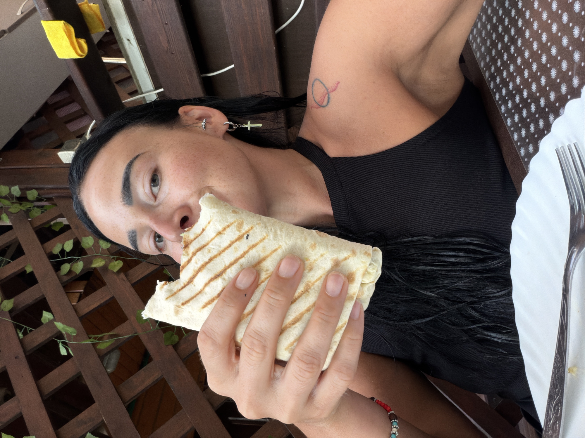 A person with long, dark hair is eating a grilled wrap. They are wearing a black tank top and silver earrings. A tattoo of an abstract oval shape is visible on their upper arm. The person is holding the wrap with their right hand, and a bite has been taken out of it. The background shows a wooden lattice structure, some greenery, and a table with a white tablecloth featuring a repeating pattern of white flowers. A fork is also visible on the table. The lighting suggests it is daytime.