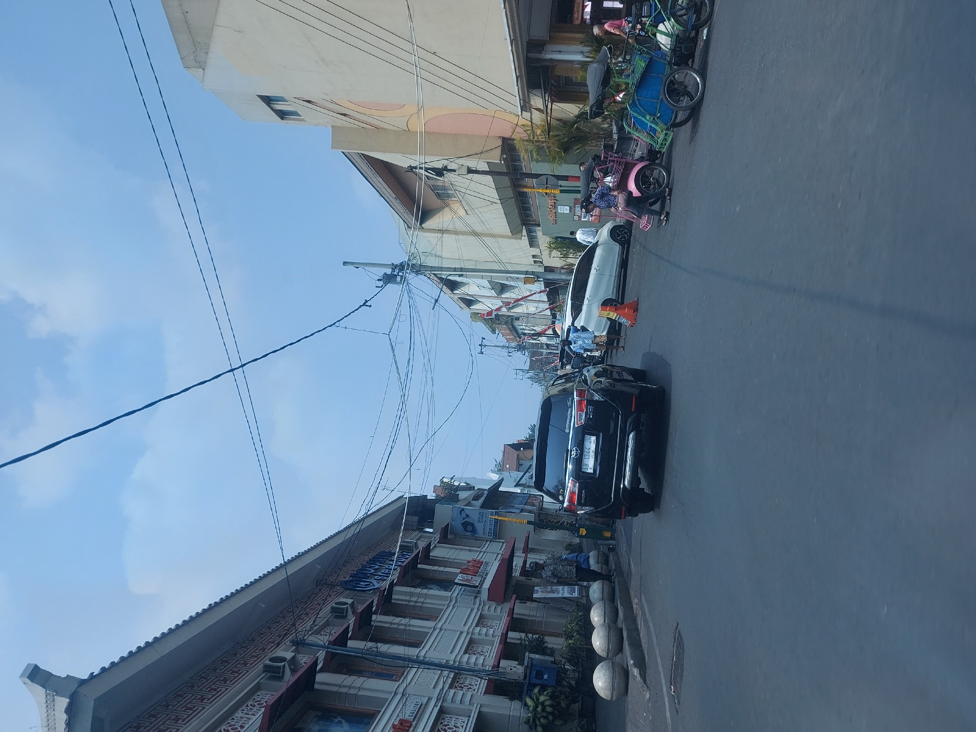 The image captures a street scene in Yogyakarta, Indonesia, likely a commercial area given the buildings lining the road. A black SUV is the dominant vehicle in the foreground, driving on the asphalt road. To the left, a row of buildings features ornate architectural details and awnings, with several air conditioning units visible. A man in a patterned shirt stands on the sidewalk next to a line of large, rounded concrete bollards. Further down the street, a white car is parked, and several people are interacting with a pink and a green pedicab. The sky above is blue with scattered clouds, and electrical wires crisscross overhead, a common sight in urban Indonesian settings. The overall impression is a bustling street on a clear day.