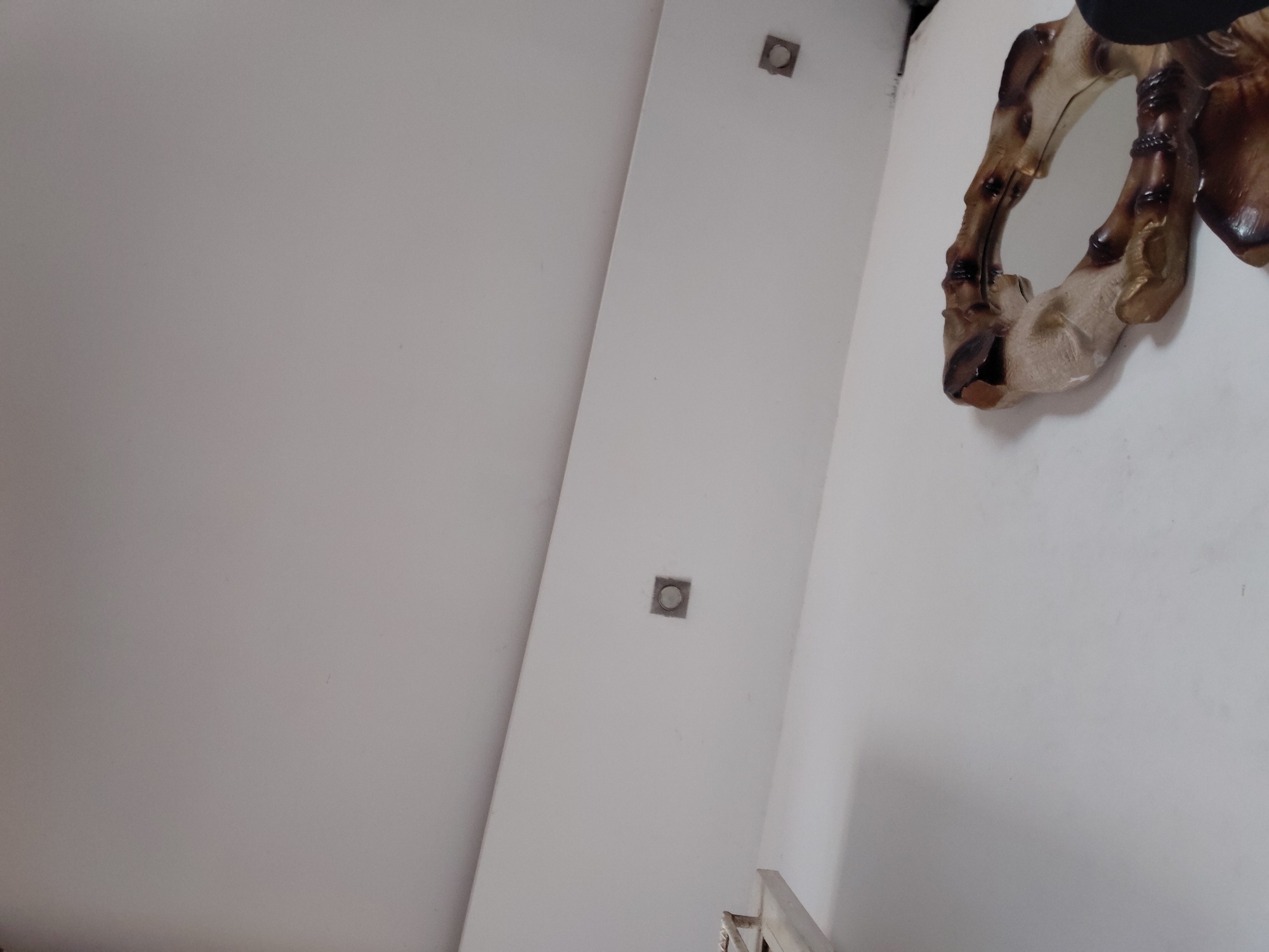 This angled indoor shot captures a section of a room or hallway, predominantly featuring light-colored walls, appearing white or very light grey, under soft, ambient illumination.
On the right side of the frame, a distinctive decorative mirror or wall art piece is mounted. Its frame exhibits an irregular, organic, or skeletal design, crafted in varying tones of brown and beige, suggesting materials like carved wood or resin. The mirror's reflective surface is partially visible but does not show clear reflections.
A prominent architectural feature, possibly a pillar or a partition, runs diagonally through the center-left of the image. Along this vertical strip, two recessed light fixtures are embedded. Each fixture has a square, metallic-looking bezel surrounding a circular light element. One is located higher up, and the other is positioned below it.
In the bottom right corner, a small portion of a light-colored structure, possibly a railing or a piece of furniture, with metallic accents, is visible. No people are present, and no specific activity is occurring. The scene conveys a quiet, static interior environment. There is no visible text in the image. The setting is an indoor space, located in Santa Ana, Venezuela.