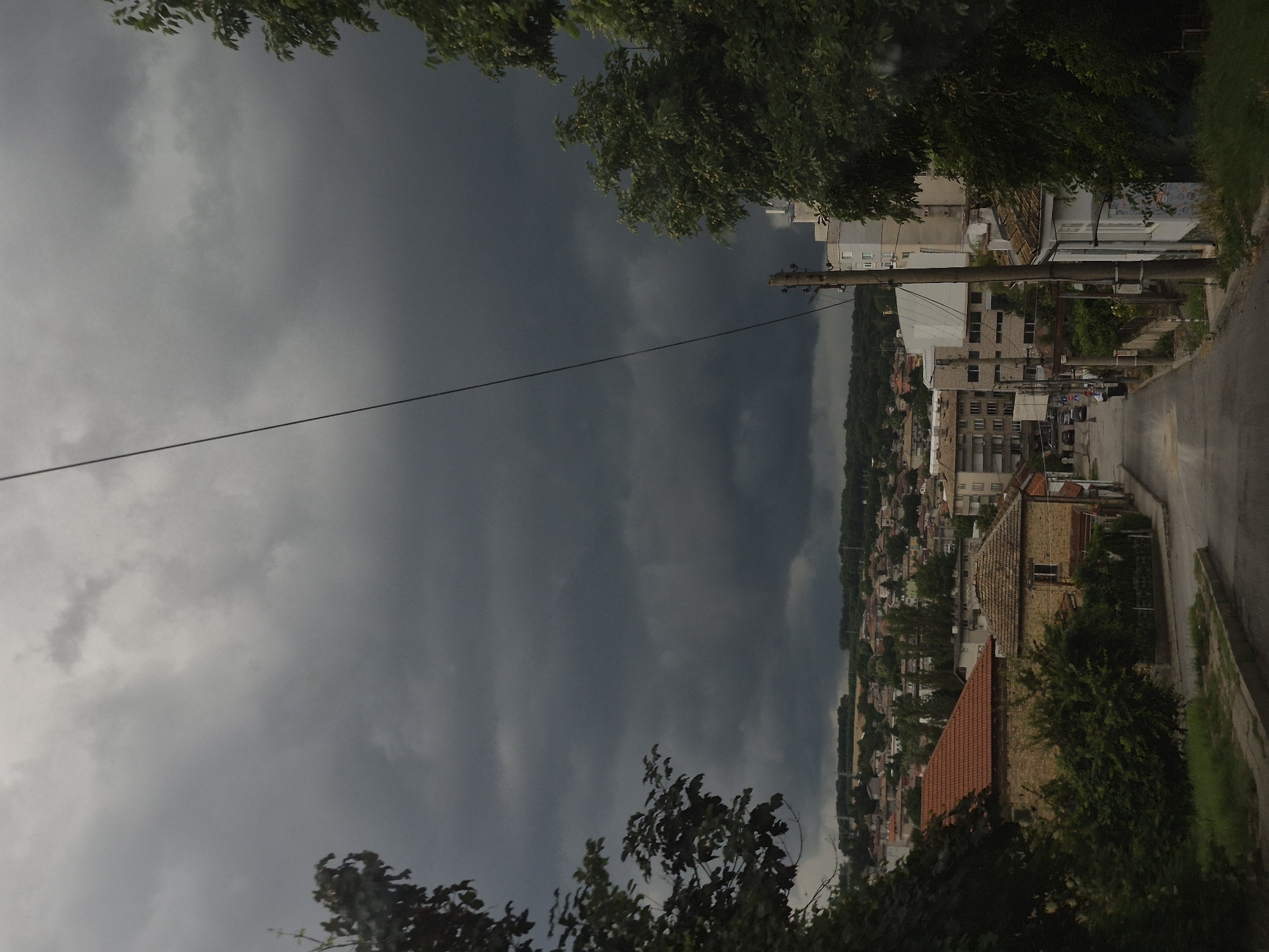 This is a photograph taken from a slightly elevated position looking down a paved street in the city of Popovo, Bulgaria. The sky is heavily overcast with dark, ominous storm clouds, suggesting an impending rain shower. On either side of the street, there are various buildings, including residential houses with red-tiled roofs, multi-story apartment buildings, and what appears to be a small commercial building with a white facade. Trees with green foliage are visible in the foreground and scattered throughout the scene, some of which are quite dense. A single utility pole with wires runs diagonally across the upper portion of the frame. The street itself is wet, indicating recent or ongoing rain. There are no people visible in the image. The overall atmosphere is somber due to the dark sky and the implied threat of a storm.