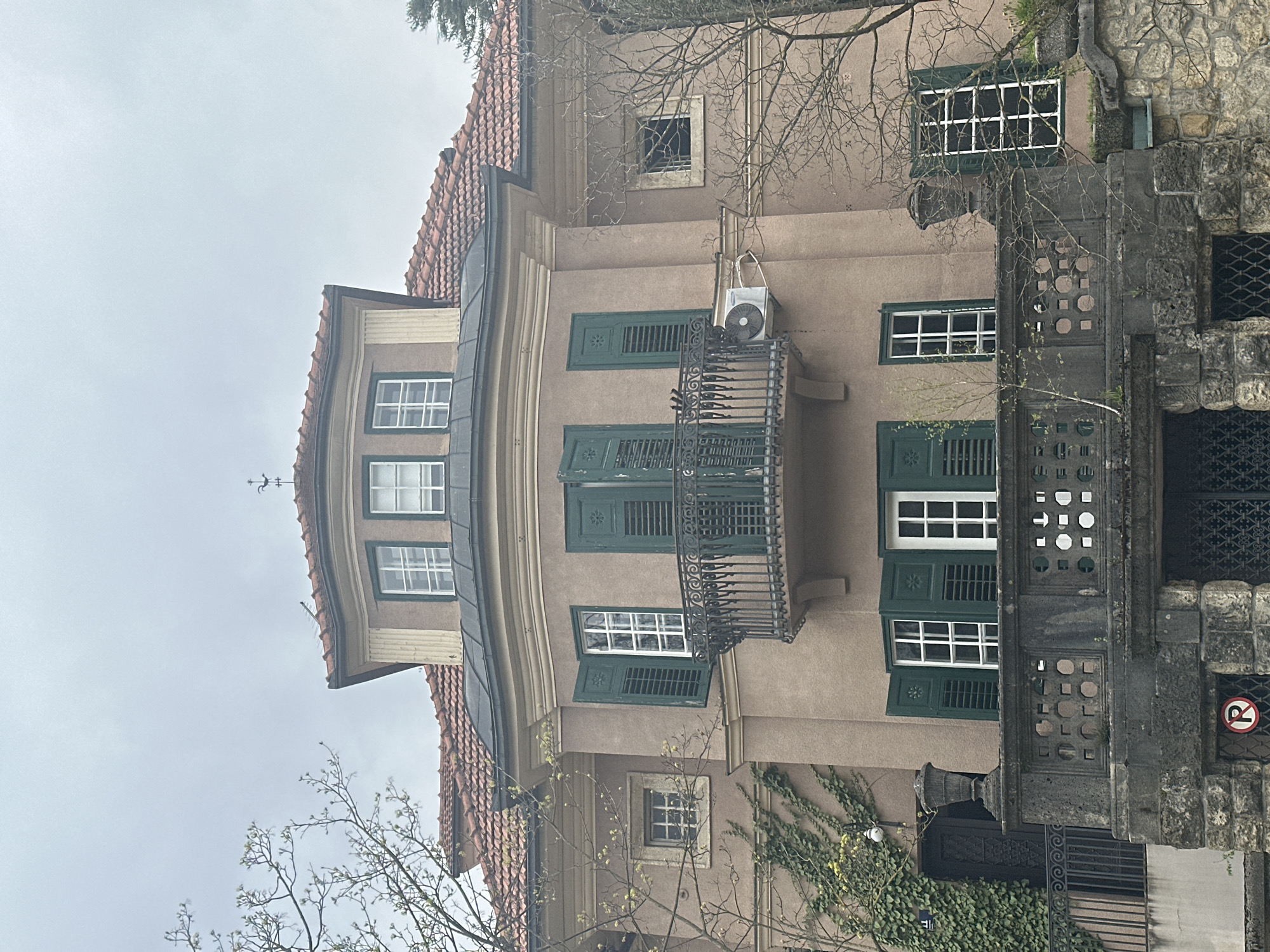The image captures a section of a building in Zagreb, Croatia, showcasing distinctive architectural features. The building, likely residential or a similar establishment, exhibits a light brown facade with a terracotta-tiled roof. A rounded projection extends from the main structure, featuring two windows on its upper level. A wrought iron balcony with dark green shutters spans the width of the main facade, above which there is an air conditioning unit. Additional windows are dispersed across the facade, some with matching dark green shutters, and one small window has no shutter. Ivy creeps up a portion of the building's exterior. Adjacent to the building, a stonework structure with ornate details and a barred window is seen, along with a "no parking" sign. Bare tree branches are visible in the foreground and above the building, indicating it may be late autumn or early spring. The sky is overcast, casting a muted light over the scene. No people are evident in the image.
