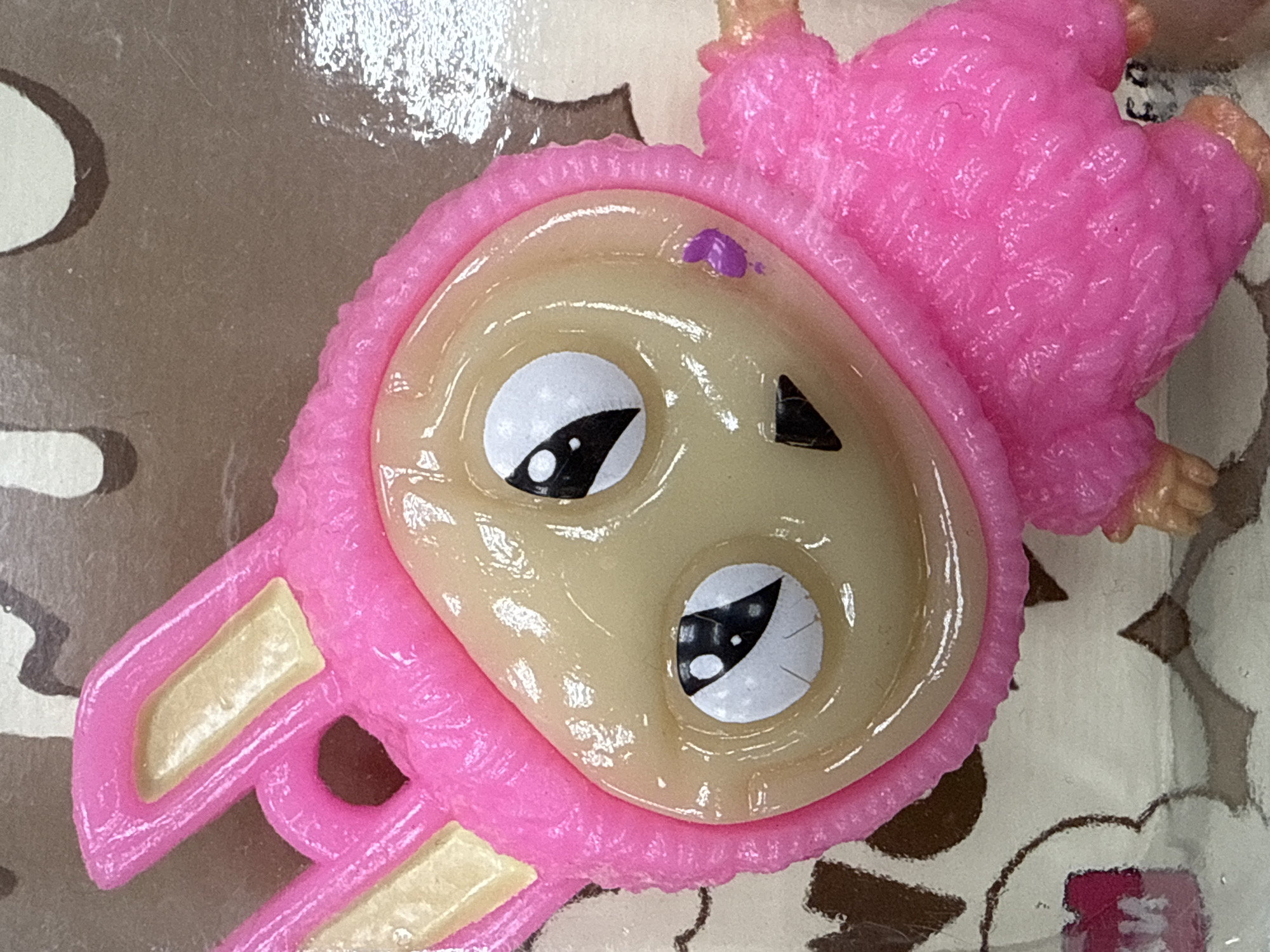 A close-up shot depicts a pink, plastic toy resembling a lamb or rabbit with a large head and stylized cartoonish eyes. The toy has a pink hood with rabbit-like ears protruding from the sides. The facial features are simplified, with large, expressive eyes and a small, triangular shape representing a nose. The body appears to be a mass of pink, textured plastic. The background appears to be a surface with a pattern or design, partially obscured. There is no indication of Votkinsk, Russia, or any specific location. No people are visible.