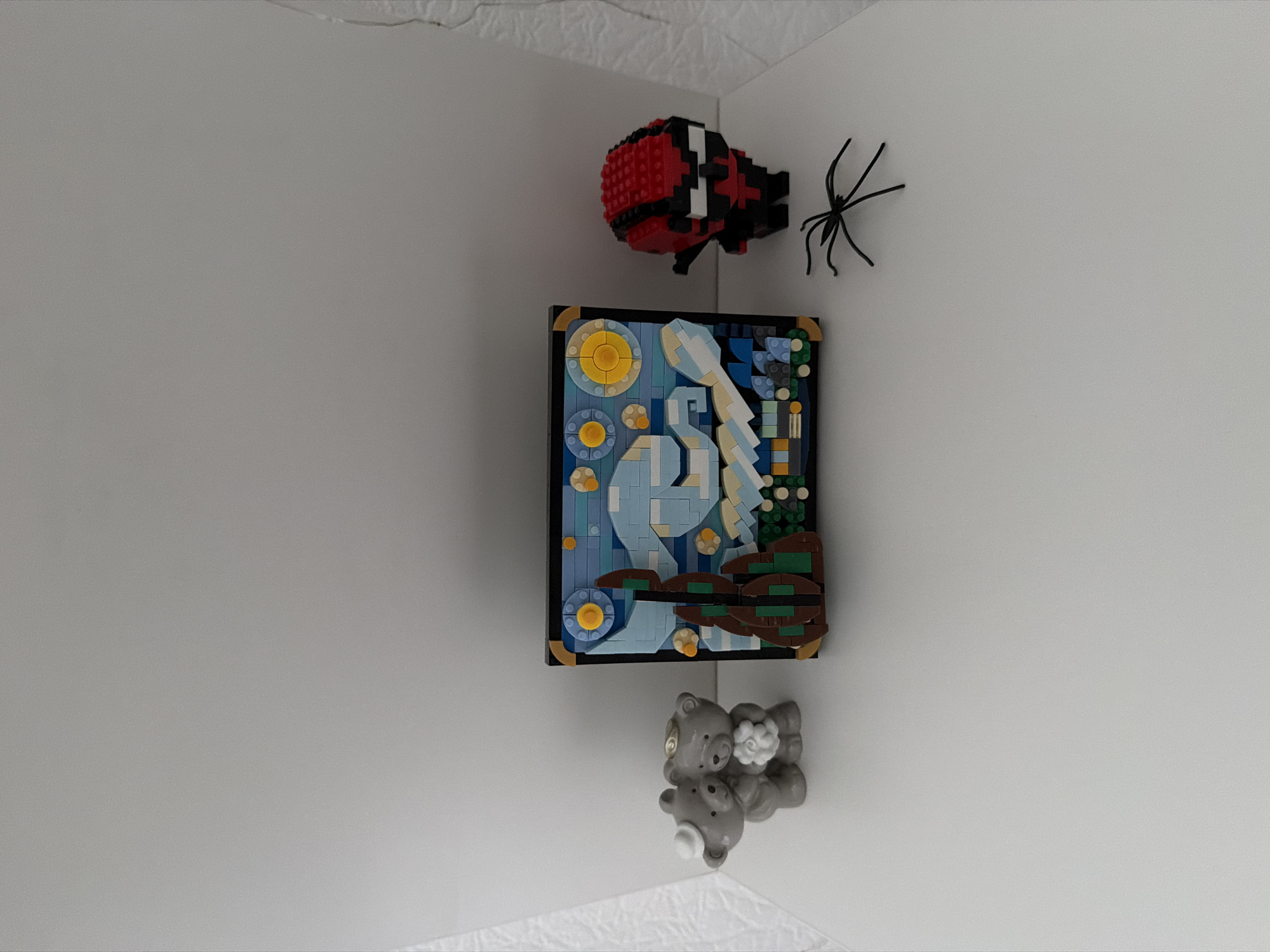 The image displays a white shelf with several items. On the left, there are two gray teddy bears embracing, with one wearing a small white hat and the other holding a white flower. To the right of the teddy bears is a framed Lego rendition of Van Gogh's "Starry Night," featuring a swirling blue sky, yellow stars, and dark cypress trees. Further to the right, a small Lego figurine with a red and black mask, resembling a superhero, stands. In front of the Lego figurine, a black plastic spider toy is positioned. The background consists of a plain white wall. The image was taken indoors, likely in a residential setting. There is no information provided about the location of this scene within the city of Kyiv, Ukraine.