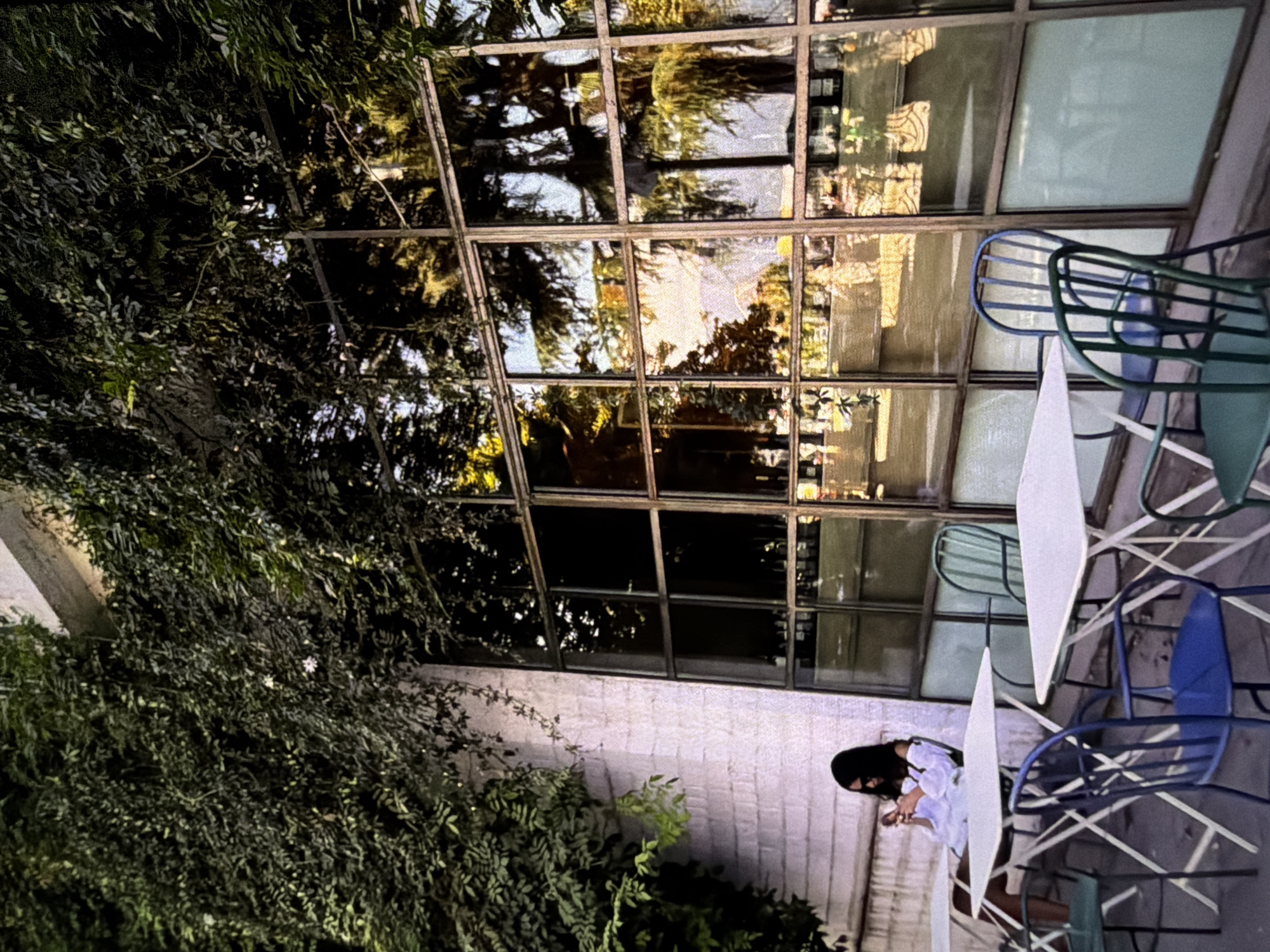 The image shows an outdoor seating area, possibly a cafe or restaurant, in an unidentified city. The seating arrangement includes square white tables surrounded by blue and green metal chairs. A person with long dark hair, dressed in white, is seated at one of the tables, appearing to be looking at a mobile phone. The area is adjacent to a building with a large grid of windows reflecting outdoor scenery, possibly trees and other buildings. Green foliage is visible on the left and at the bottom of the frame, suggesting that the area is partially shaded by vegetation.