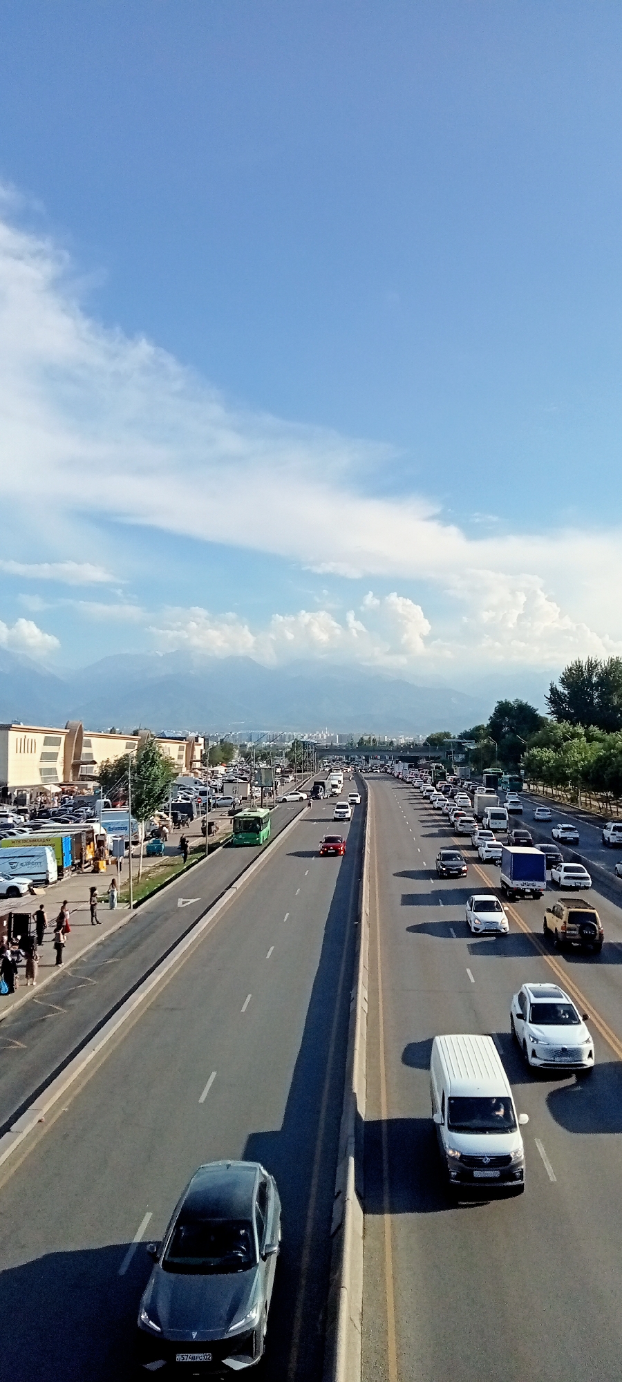 The image shows a busy multi-lane highway in Almaty, Kazakhstan, with a line of cars and trucks. The highway is bordered by a sidewalk with pedestrians and parked vehicles on the left side, and a grassy area with trees on the right. In the background, there are buildings and distant mountains under a partly cloudy sky. The scene appears to be a typical urban traffic situation with a mix of commercial and private vehicles.