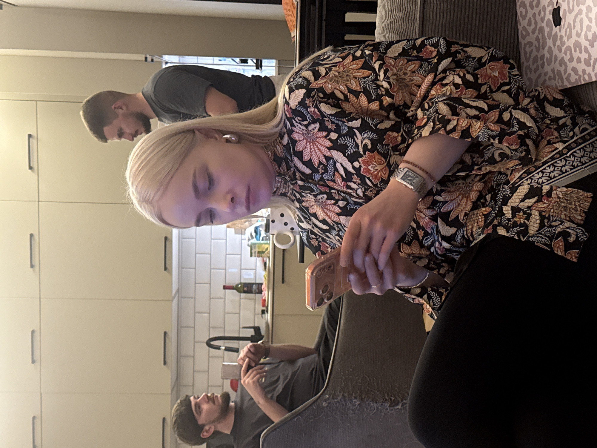 A horizontal medium shot depicts a woman with blonde hair looking down at a phone in her hands. She wears a black floral-patterned top and black pants. A man in a gray shirt stands behind her to the left, and another man in a gray shirt sits in the background near the kitchen. The background features light-colored cabinets, a white tiled wall, and a sink with a black faucet. The woman is seated on a gray couch. The scene appears to be indoors, likely in an apartment. While the location is in Batumi, Georgia, this cannot be discerned from the image alone. There are no specific indicators to suggest it.