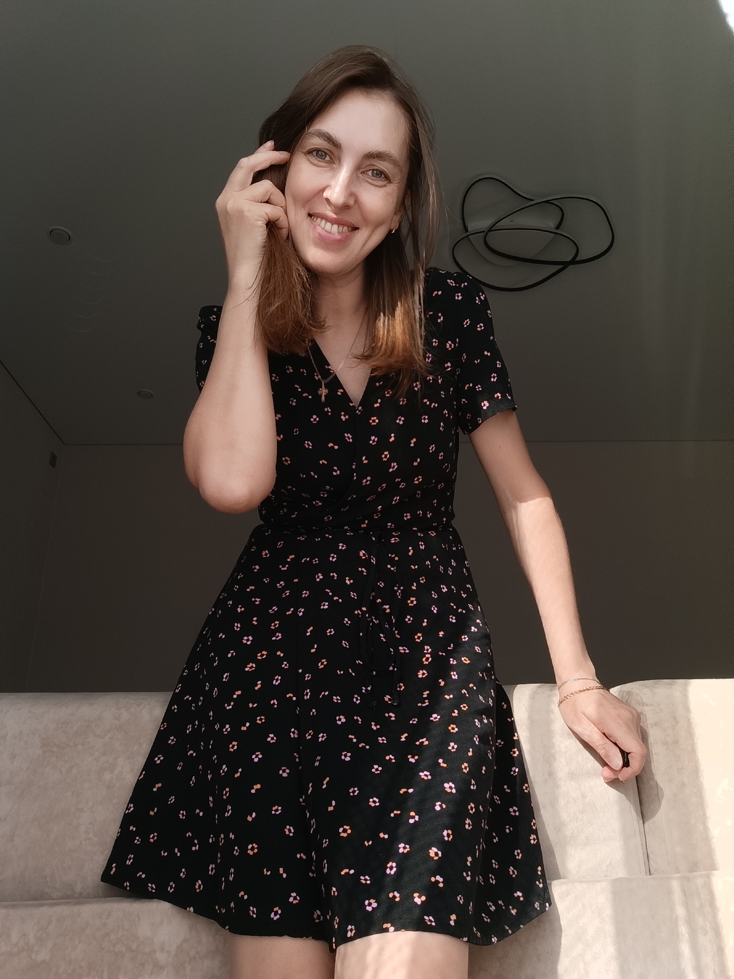 A woman is photographed indoors during daylight in what is likely an apartment. She is fair-skinned with shoulder-length brown hair and is wearing a black dress with a pattern of small, stylized flowers in orange and pink. She is smiling and has her right hand near her face, appearing to playfully touch her hair. Her left arm rests on what appears to be the armrest of a couch or similar upholstered furniture. The background is a lightly lit interior space with a minimalist aesthetic. A modern light fixture is visible on the ceiling. The image is taken in Chaikovsky, Russia.
