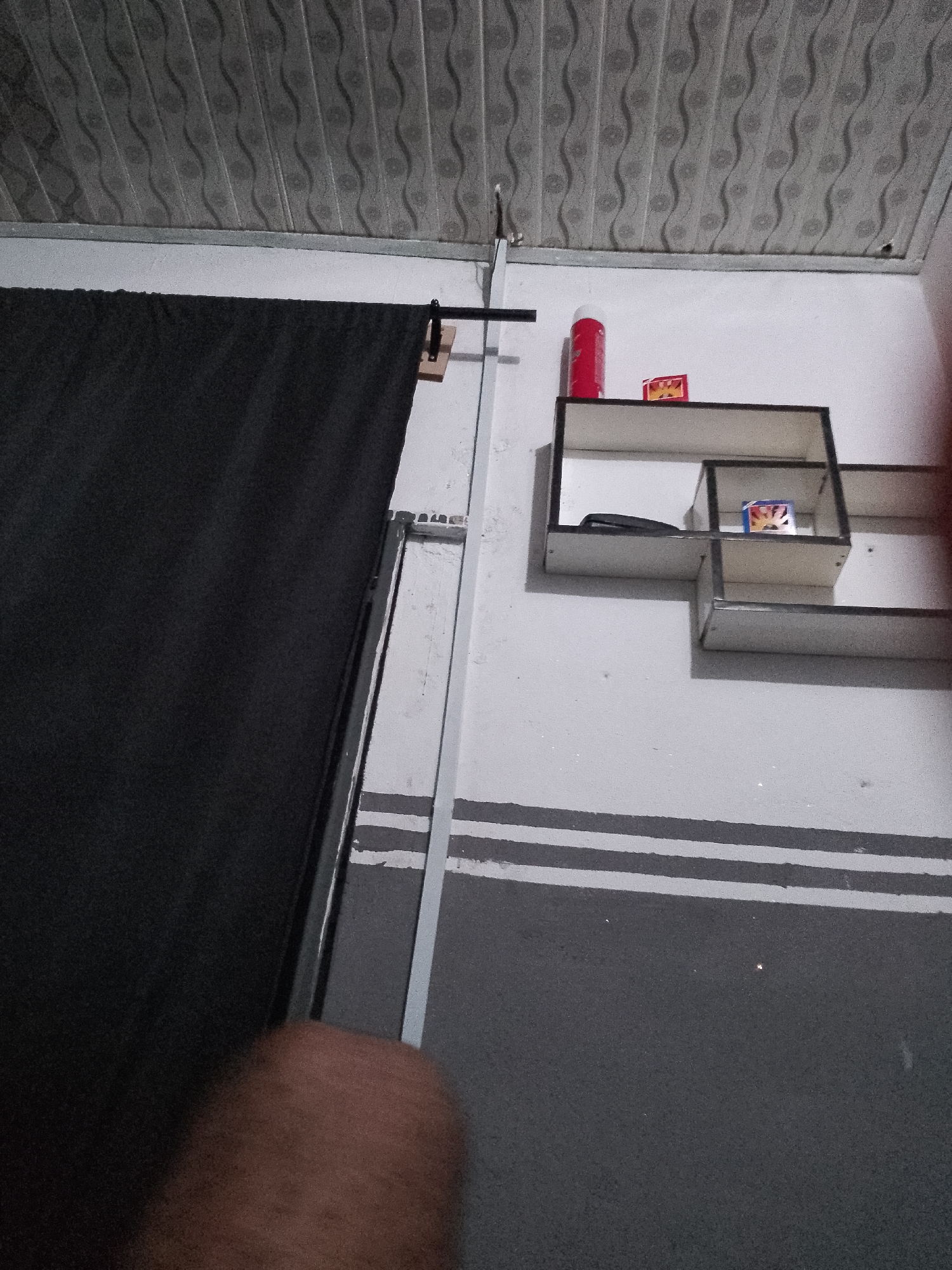 The image depicts an indoor scene. A black curtain hangs on the left side, supported by a rod attached to a wall. The wall has a white surface with some horizontal grey stripes painted on the lower half. Above the wall is a textured ceiling. On the right side of the wall, there is a set of floating shelves. On the top shelf, there is a red cylindrical object, possibly a can, and a small yellow box. On a lower shelf, there is a black object that appears to be a phone or a small electronic device, and another small box with a colorful illustration. A vertical metal pole runs from the ceiling down the wall, to the left of the shelves. A person's hand is partially visible in the lower left corner of the image. The overall impression is of a simple, utilitarian space. There is no information in the image to indicate its location in Jauro Yinu, Nigeria.