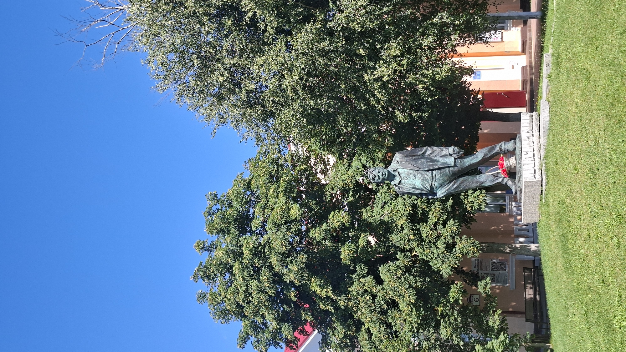 The photograph captures a bronze statue of a man in a suit standing on a stone pedestal. The statue is situated in a park-like setting with a bright blue sky overhead. Two large, leafy trees frame the statue, one on the left and another on the right. The tree on the left appears to be a horse chestnut tree, judging by its distinctive leaves and the small, spiky fruit visible on its branches. In front of the statue, a small woven basket filled with red flowers rests on the pedestal. To the left of the statue, a wooden bench sits on a patch of grass. Behind the bench, a building with a red roof and an address number "6" is visible. In the background, the building's facade is painted in warm, muted tones. The overall scene suggests a public space, possibly a memorial or a monument in the town of Myadzyel, Belarus.
