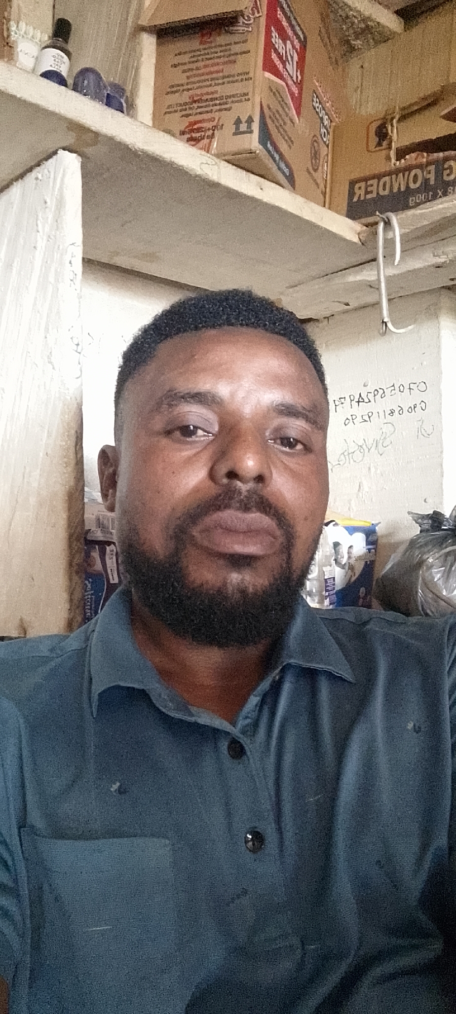 A man is pictured from the chest up, looking directly at the camera. He has dark skin, short, dark, curly hair, and a full beard and mustache. He is wearing a dark blue collared shirt. The background appears to be a shelf or storage area, with various items on it, including boxes and what looks like a roll of paper towels or toilet paper. Some writing is visible on the wall in the background. The lighting suggests an indoor setting, possibly a shop or storage room. The man appears to be in an urban environment, and based on the request to include location details, this scene could be in the city of Durumi, Nigeria.