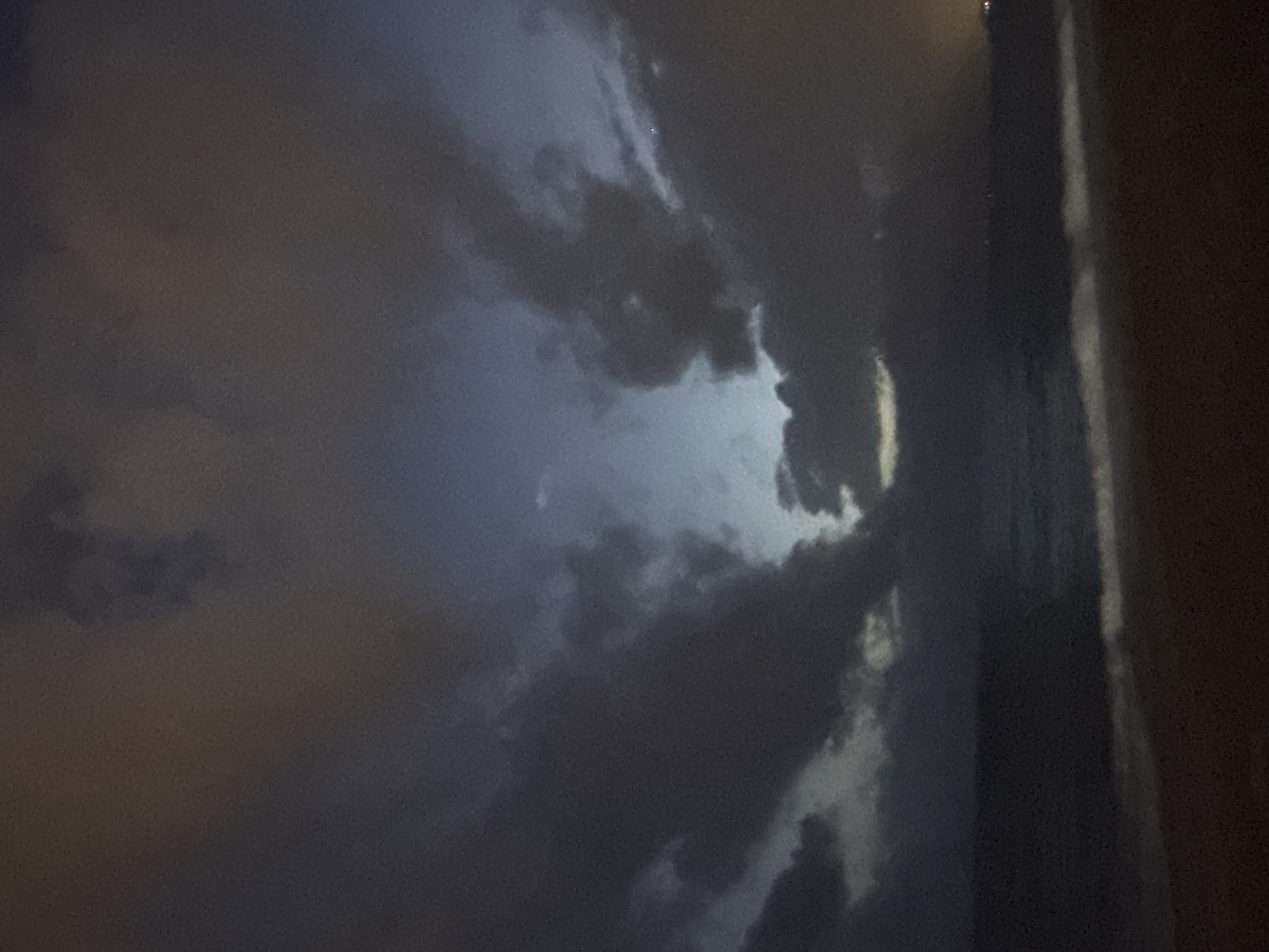 The image shows a dark, cloudy sky at night. The scene is likely in Alanya, Turkey. The clouds are thick and appear ominous, with a possible break in the clouds revealing a lighter area or the moon. The right side of the image shows a dark, solid surface, perhaps a building or wall. The overall mood is one of a stormy or unsettled weather condition. There is no visible sign of people or action.