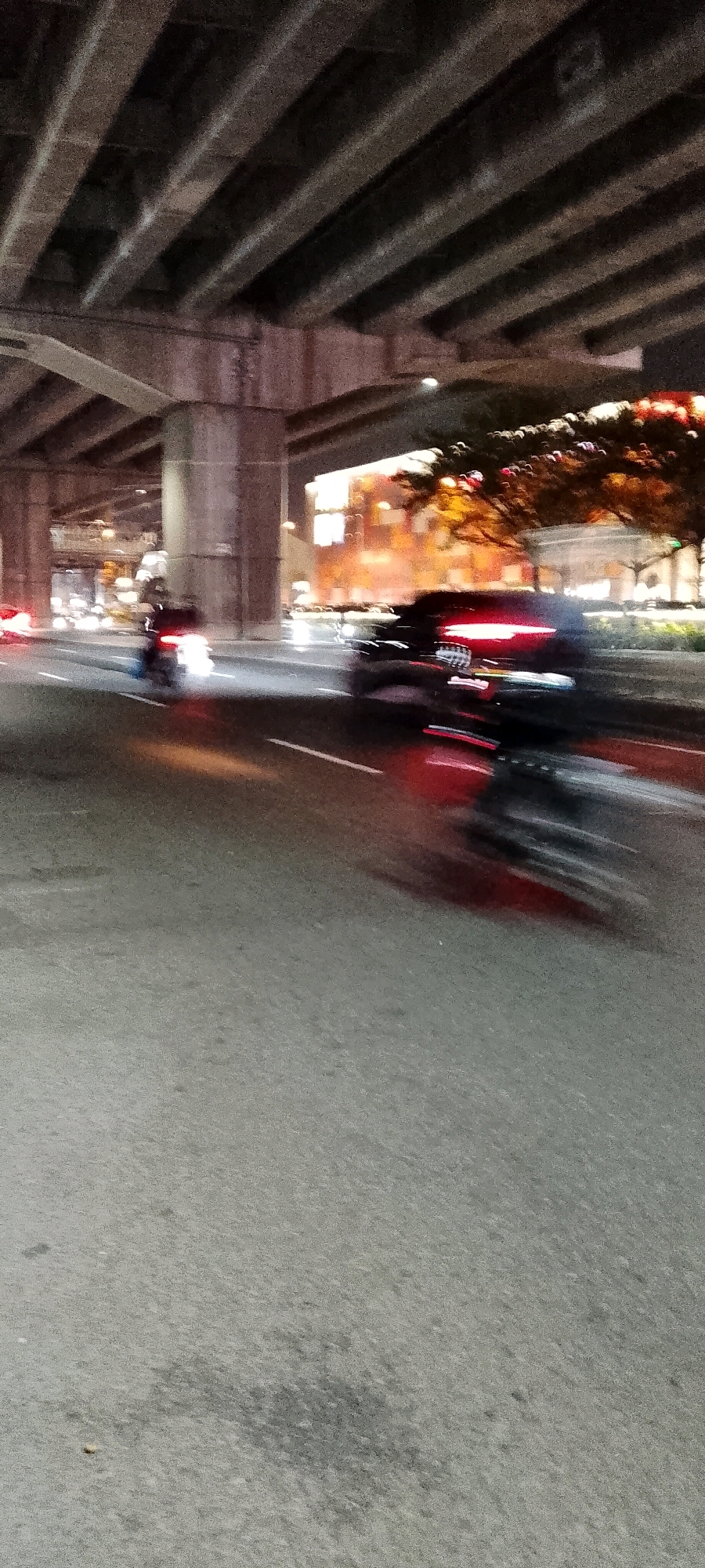The image captures a nighttime street scene in Bekasi, Indonesia, characterized by motion blur due to a slow shutter speed. The foreground is dominated by a gray asphalt road, showing some wear and patches of moisture. Above the road, a concrete overpass with parallel beams stretches across the frame, casting shadows and defining the upper boundary of the scene.
On the road, several vehicles are in motion. A dark-colored car, likely an SUV, is prominently featured in the middle ground, its red taillights streaking across the image. To the left, a motorcycle is visible, its headlight creating a bright white blur. Further in the distance, a line of blurred vehicle lights suggests ongoing traffic.
The background shows illuminated buildings, possibly storefronts or residences, with warm orange and yellow lights. Trees with autumn foliage are also visible, their colors muted by the darkness and motion blur. The overall impression is one of a busy urban environment at night, with the overpass adding a sense of depth and scale to the scene.