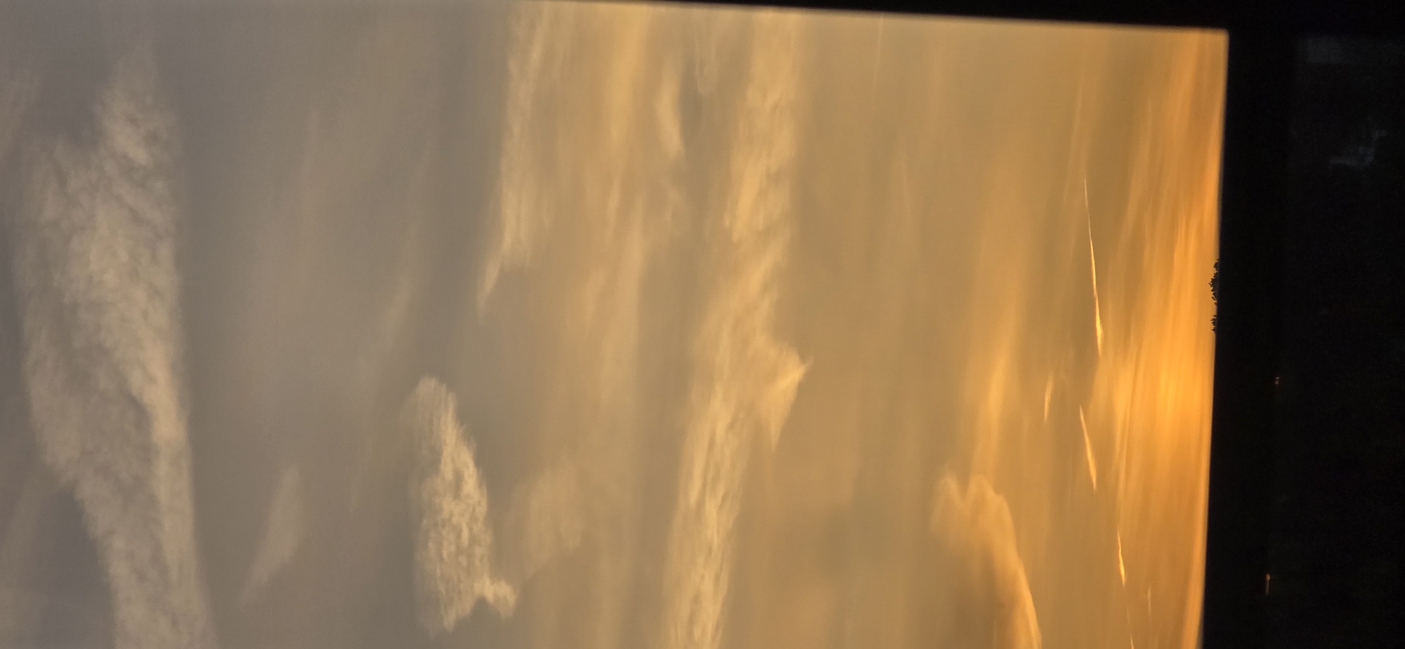 The image shows a sunset over an urban silhouette. The sky is a gradient of orange, yellow, and gray, with wispy clouds streaked across it. The sun is low on the horizon, casting a warm glow. Below the sky, a dark, silhouetted line of buildings and trees stretches across the frame, suggesting the presence of a city. The foreground is also dark, indicating the viewer is likely indoors looking out a window, with a dark frame visible on the right side of the image. There are no people or specific objects identifiable in the scene.