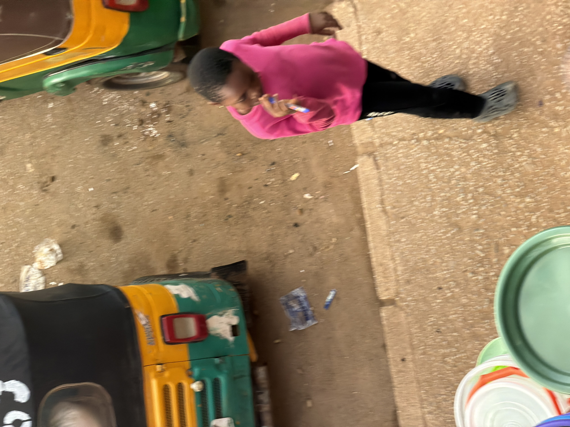An overhead shot captures a young girl in a bright pink long-sleeved shirt and black leggings, standing on a sandy, rough textured surface. She is holding a blue and white object, possibly a pen or crayon, in her right hand, and appears to be in motion. To her left, partially visible, is the back of a yellow, green, and black auto-rickshaw, which is parked on the same surface. Further to the left, a collection of colorful plastic buckets and lids is stacked. The ground is littered with small debris, including what appears to be crumpled white paper and a thin blue object. The overall scene suggests an outdoor setting in a casual, possibly urban or semi-urban environment. The question mentions Bukuru, Nigeria, but this cannot be confirmed from the image alone.