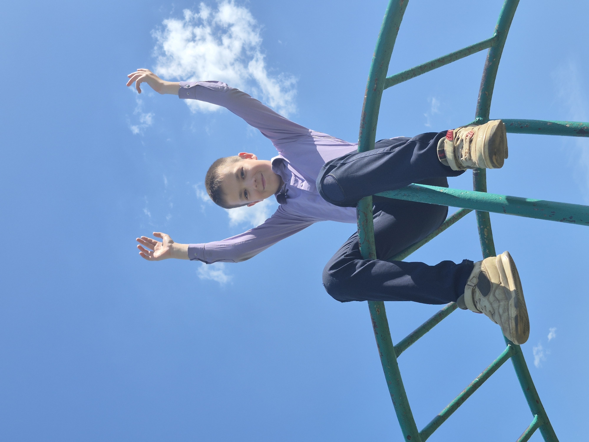 A boy in a long-sleeved light purple collared shirt and dark blue pants is hanging upside down from a green metal playground structure against a clear blue sky with scattered white clouds. He is wearing tan and red sneakers and has his arms extended out to the sides. The location is the city of Chaikovsky, Russia.