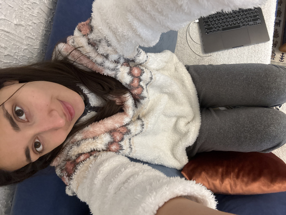 This image depicts a female individual in an indoor setting, identified as Zankovići, Montenegro. The subject is seen from an overhead perspective, looking directly at the camera with a slight smile. She has dark hair, dark eyes, and a light complexion. She is wearing a cream-colored, fluffy pullover top featuring a patterned design in pink, white, and dark gray around the chest and shoulders, and dark gray trousers. To her left, a dark blue sofa or cushion is partially visible, with an orange-brown velvet-textured cushion resting against it. The floor in the foreground is covered by a light-colored, textured rug. A dark gray laptop with a QWERTY keyboard is positioned on this rug to the subject's right, connected by a white charging cable. A section of a dark patterned rug is visible beneath the laptop at the bottom right. The background on the upper right displays a light-colored, textured wall.